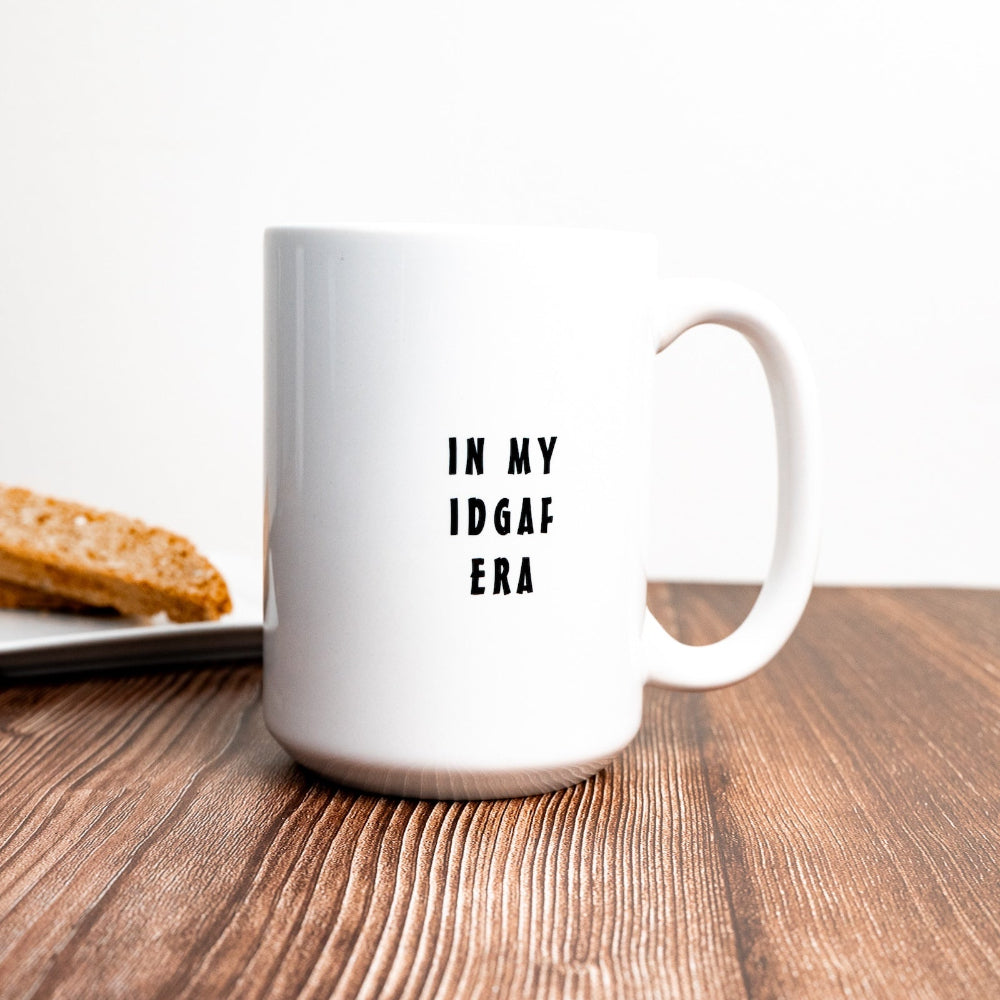 A white ceramic mug with the text 'IN MY IDGAF ERA' printed in black color, placed on a wooden surface with a piece of bread in the background.