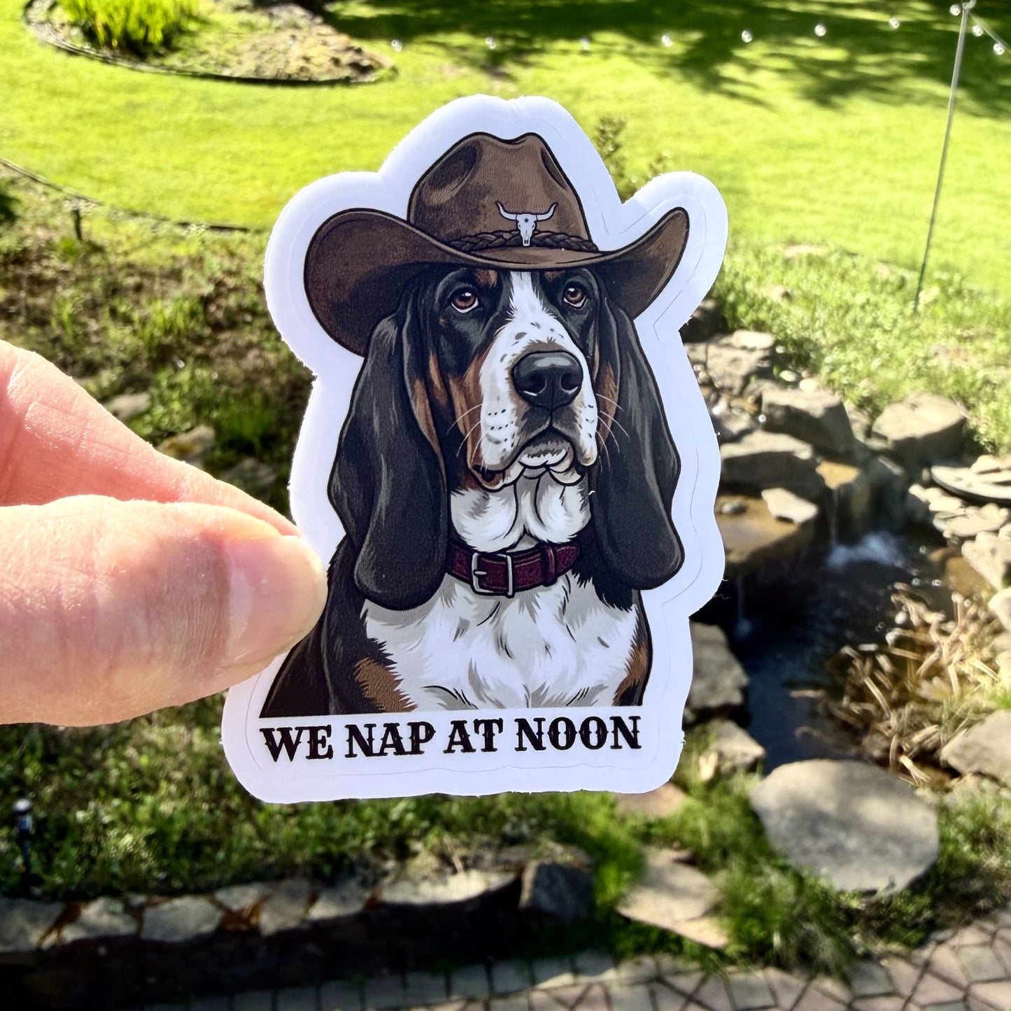 Cowboy basset hound sticker - dog wearing a cowboy hat with 'WE NAP AT NOON' text, held in front of a garden background.