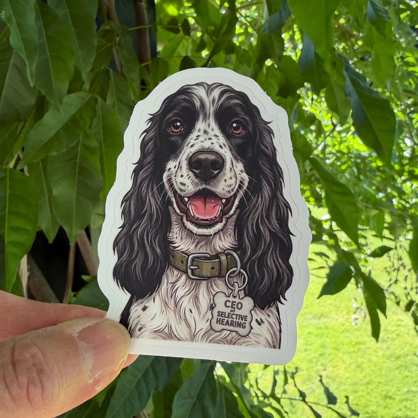 Cute English Cocker Spaniel sticker held in one hand against leafy green trees, showing the illustrated black-and-white Spaniel portrait and white kiss-cut border.