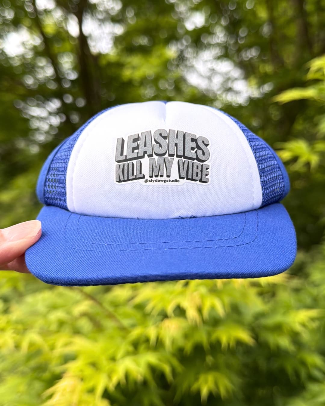 Dog trucker hat with saying lifestyle blue; blue and white Leashes Kill My Vibe dog trucker hat held outdoors with green foliage in the background.