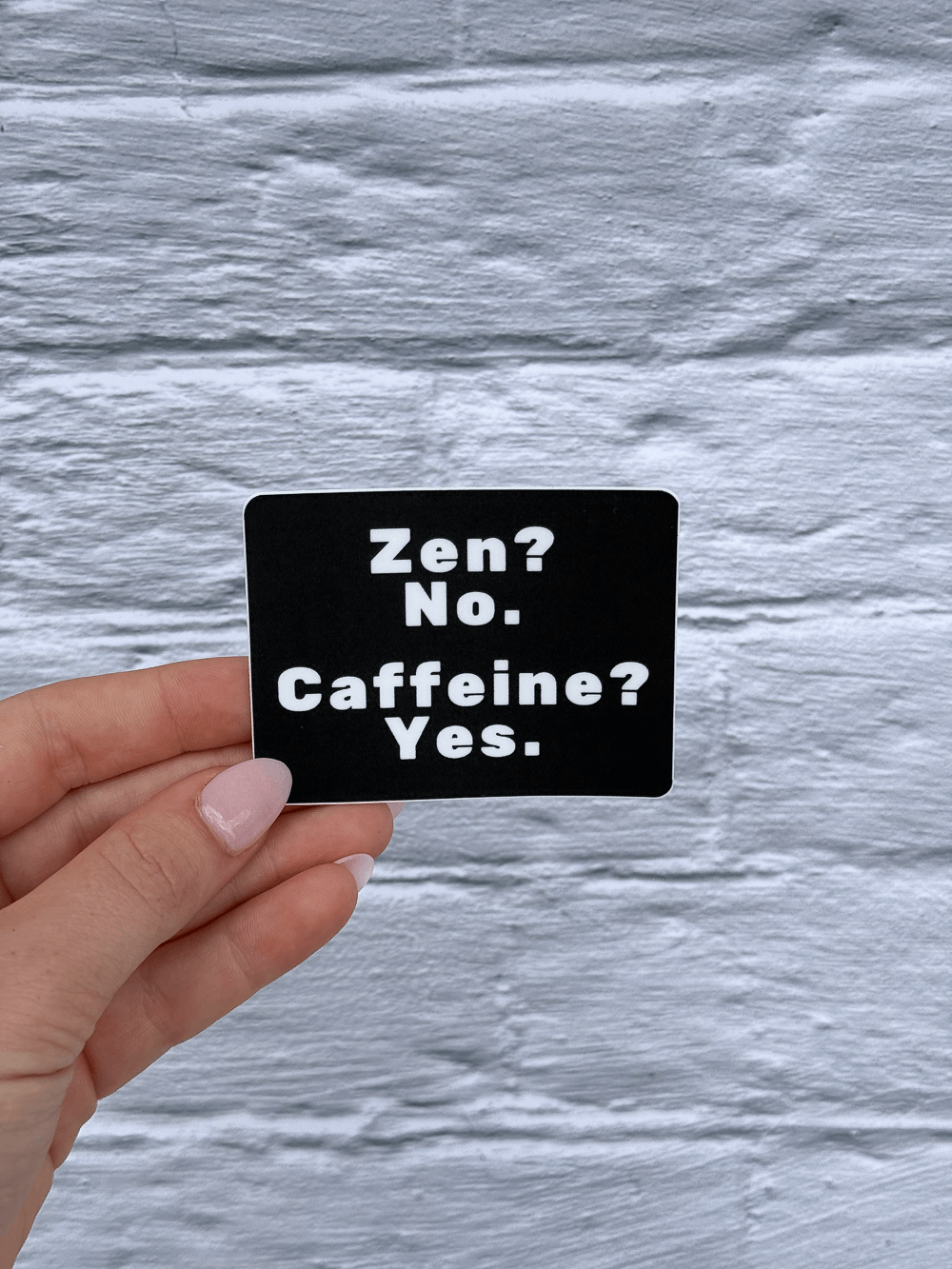 Hand holding a funny caffeine sticker with white text against a gray textured background