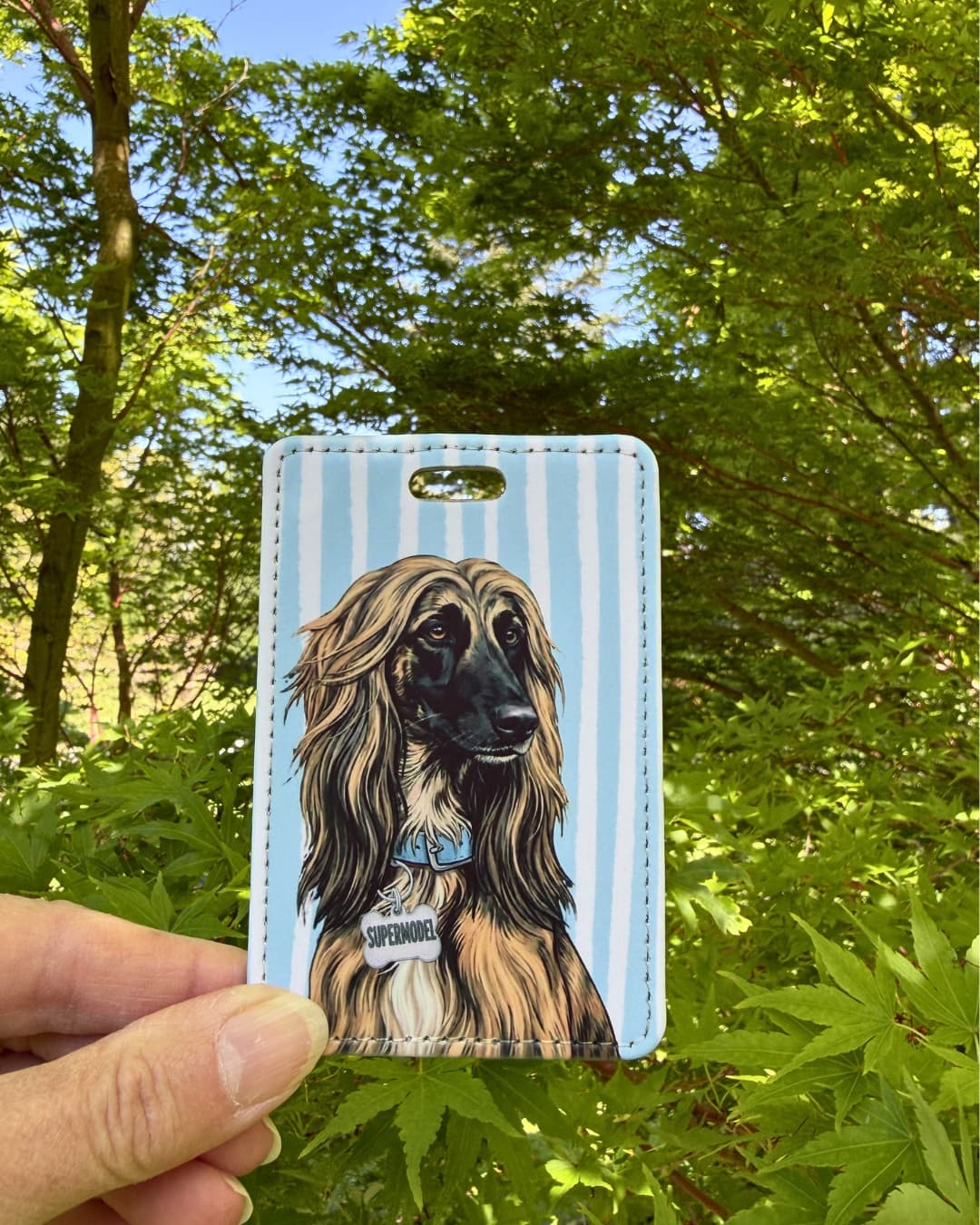Afghan hound luggage tag lifestyle; Hand holding a fun faux leather luggage tag featuring an Afghan Hound with flowing tan hair and a Supermodel charm, vertical light blue and white stripes, green tree foliage background.