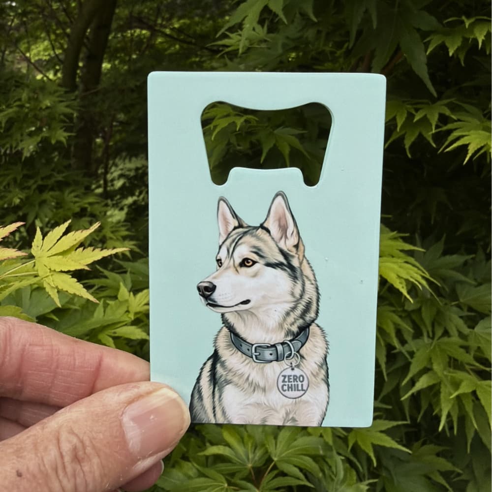 Agouti husky bottle opener:  A hand holding a credit card sized agouti husky bottle opener outdoors with green leaves in the background.