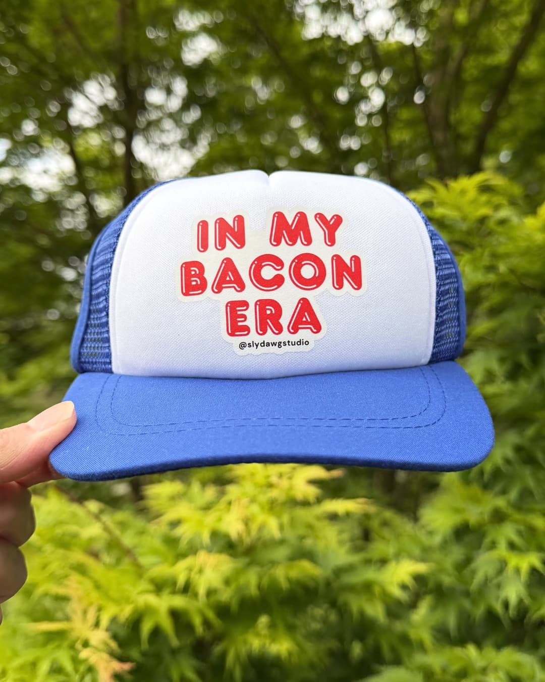 Bacon lover dogs hat lifestyle; hand holding a blue and white In My Bacon Era dog trucker hat outdoors with green Japanese Maple leaves in the background.