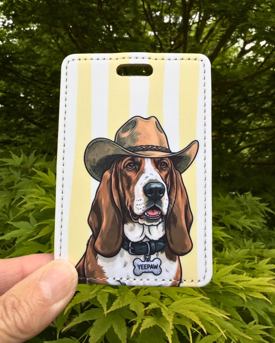 Basset Hound cowboy luggage tag lifestyle; hand holding the faux leather Basset Hound Yeepaw luggage tag outdoors with green Japanese Maple leaves in the background.