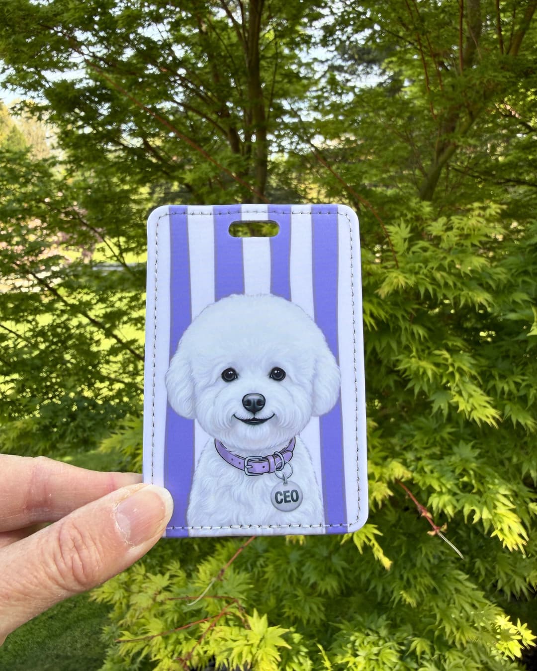 Bichon Frise luggage tag lifestyle; Hand holding a fun faux leather luggage tag featuring a fluffy white Bichon Frise with a purple collar and CEO charm, vertical purple and white stripes, green tree foliage background.