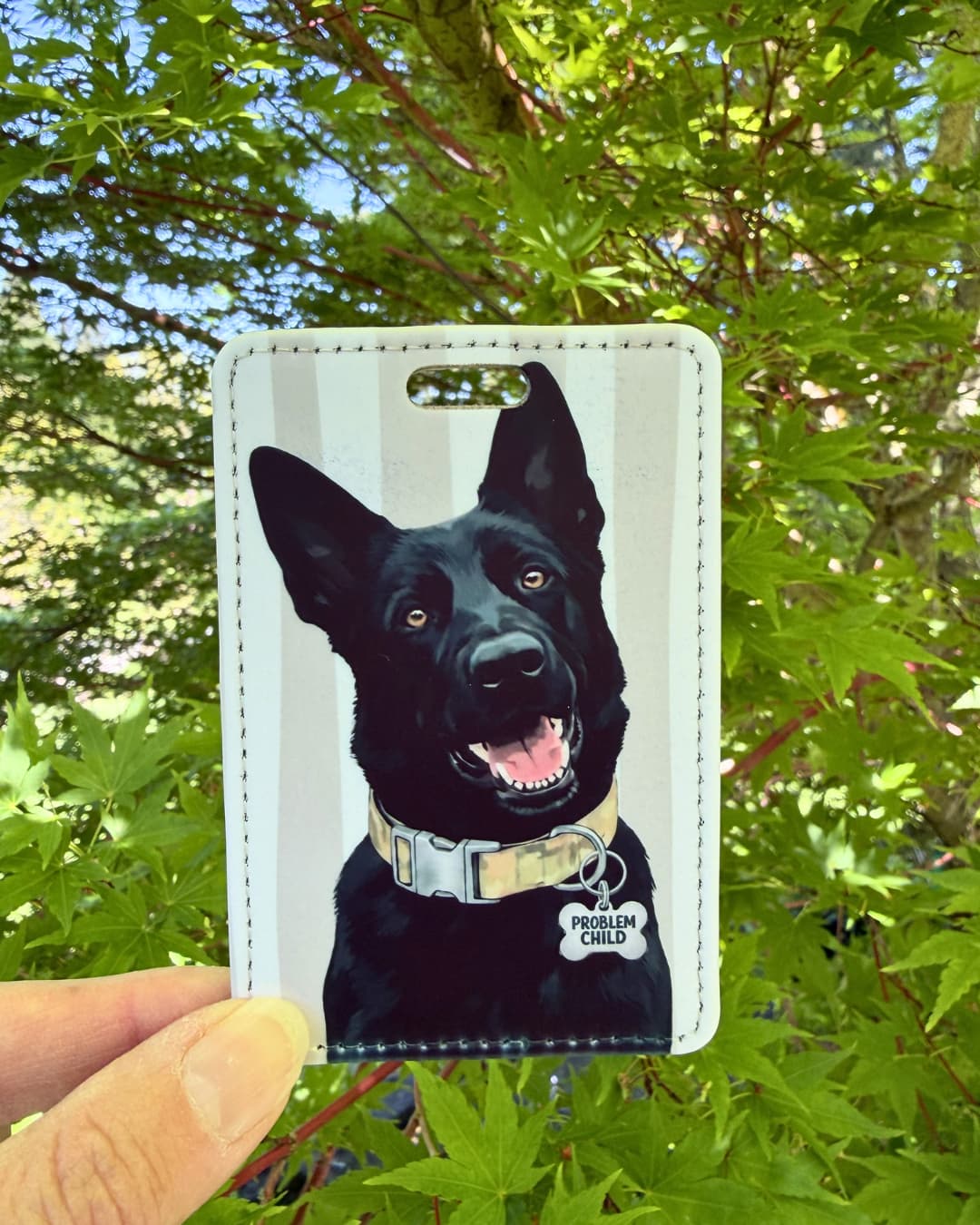 Black German Shepherd luggage tag lifestyle; Hand holding a vibrant faux leather luggage tag featuring a grinning black GSD with a Problem Child funny bone charm, grey and white stripes, green tree foliage background.