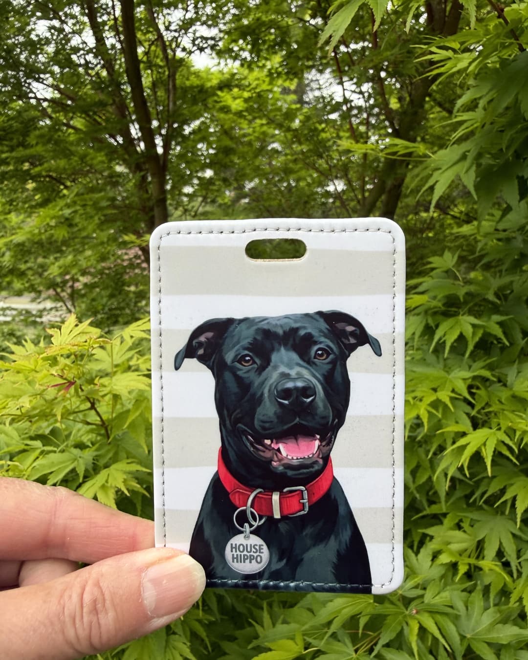 black pitbull luggage tag handheld outdoors against green foliage showing smiling black Pitbull with red collar and HOUSE HIPPO collar tag on taupe and white striped faux leather tag