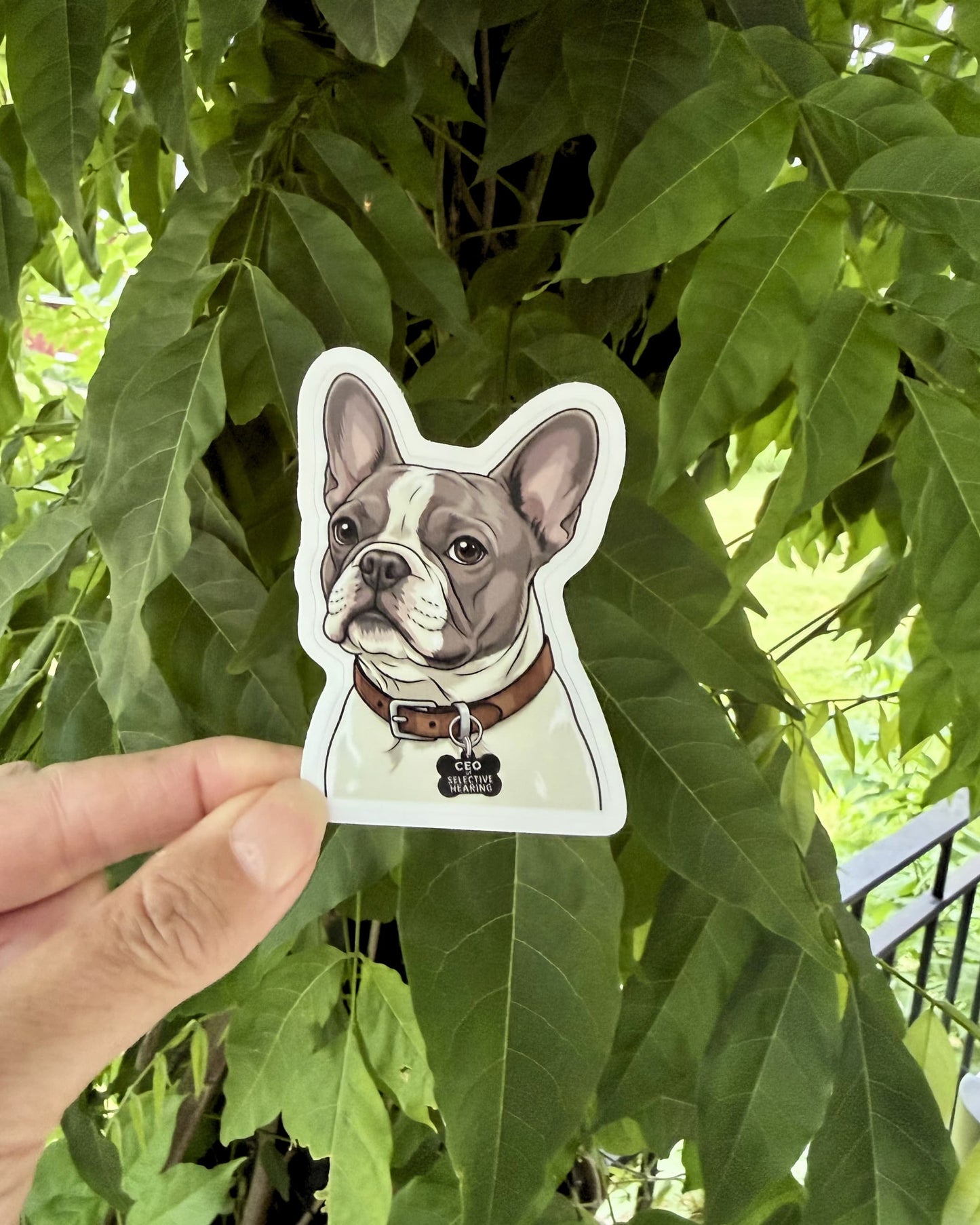 Hand holding blue pied French Bulldog waterproof kiss-cut sticker outdoors against green leaves.