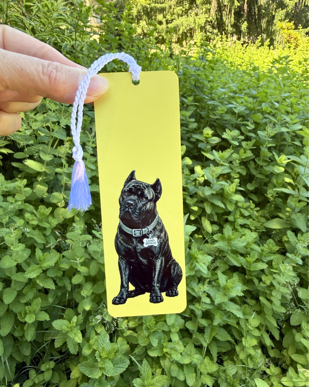 Hand holding brindle Cane Corso bookmark against green foliage, golden yellow aluminum, Land Whale collar tag, white tassel