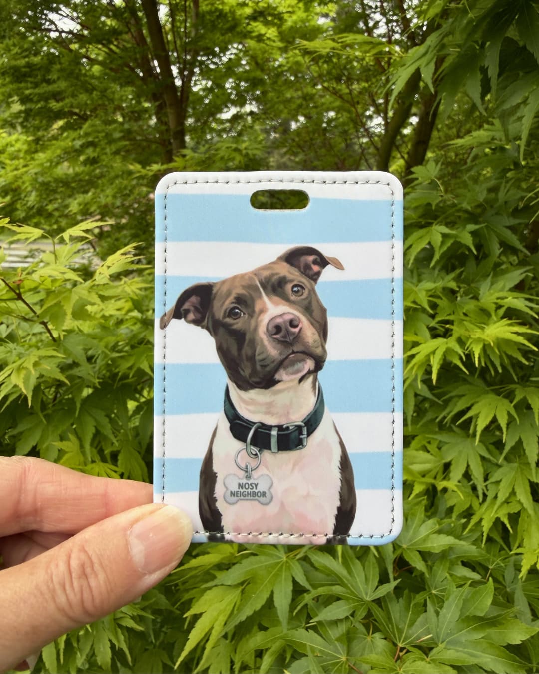 Brown and white pitbull luggage tag lifestyle; Hand holding a fun faux leather luggage tag featuring an illustrated brown and white Pitbull with a Nosy Neighbor charm, light blue and white horizontal stripes, green tree foliage background.