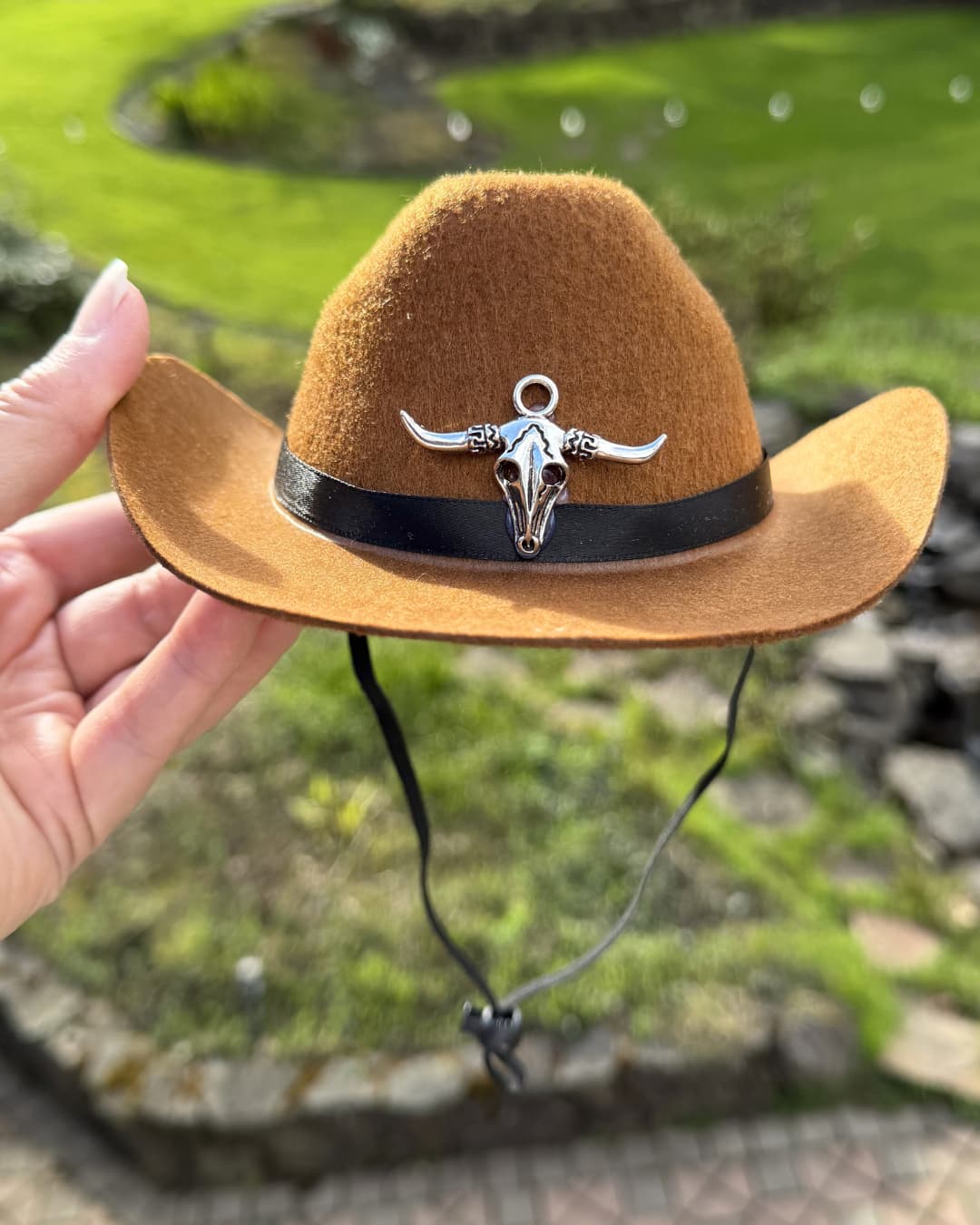 Brown cowboy hat for dogs with a bull skull charm held by a hand against a natural background.