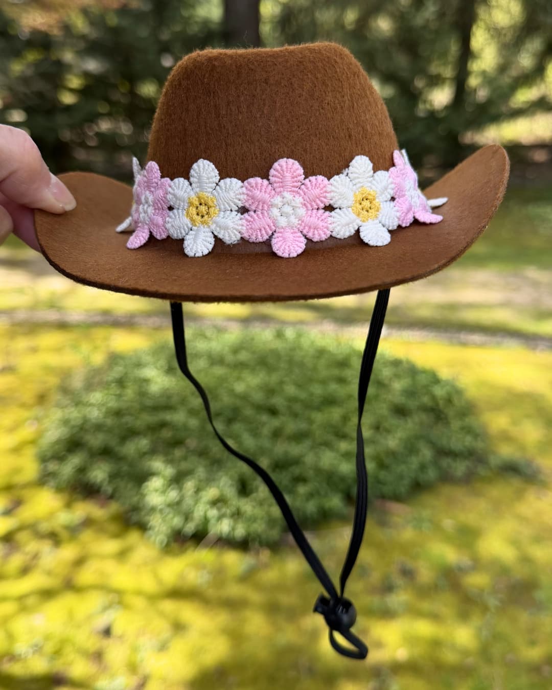 Brown floral daisy cowboy hat with a floral headband held against a blurred natural background