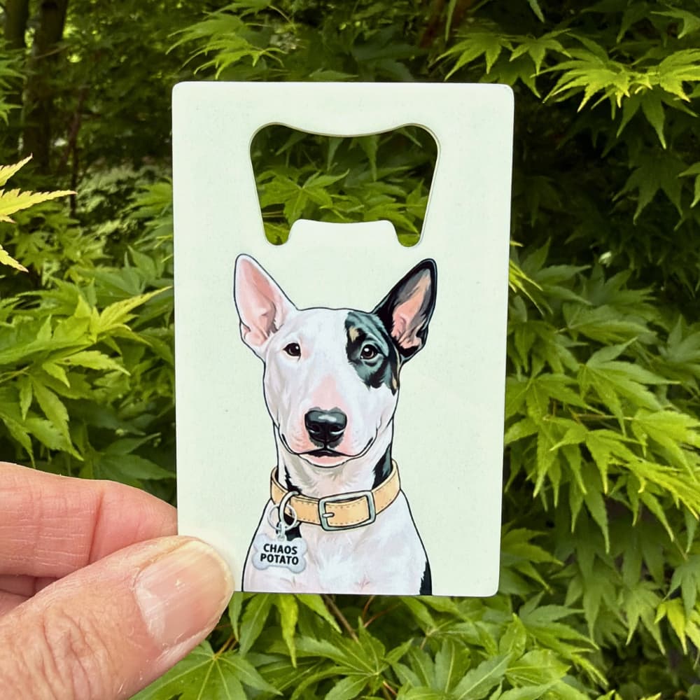 Hand holding a credit card sized Bull Terrier bottle opener outdoors with green Japanese Maple leaves in the background - Chaos Potato bully portrait on stainless steel.
