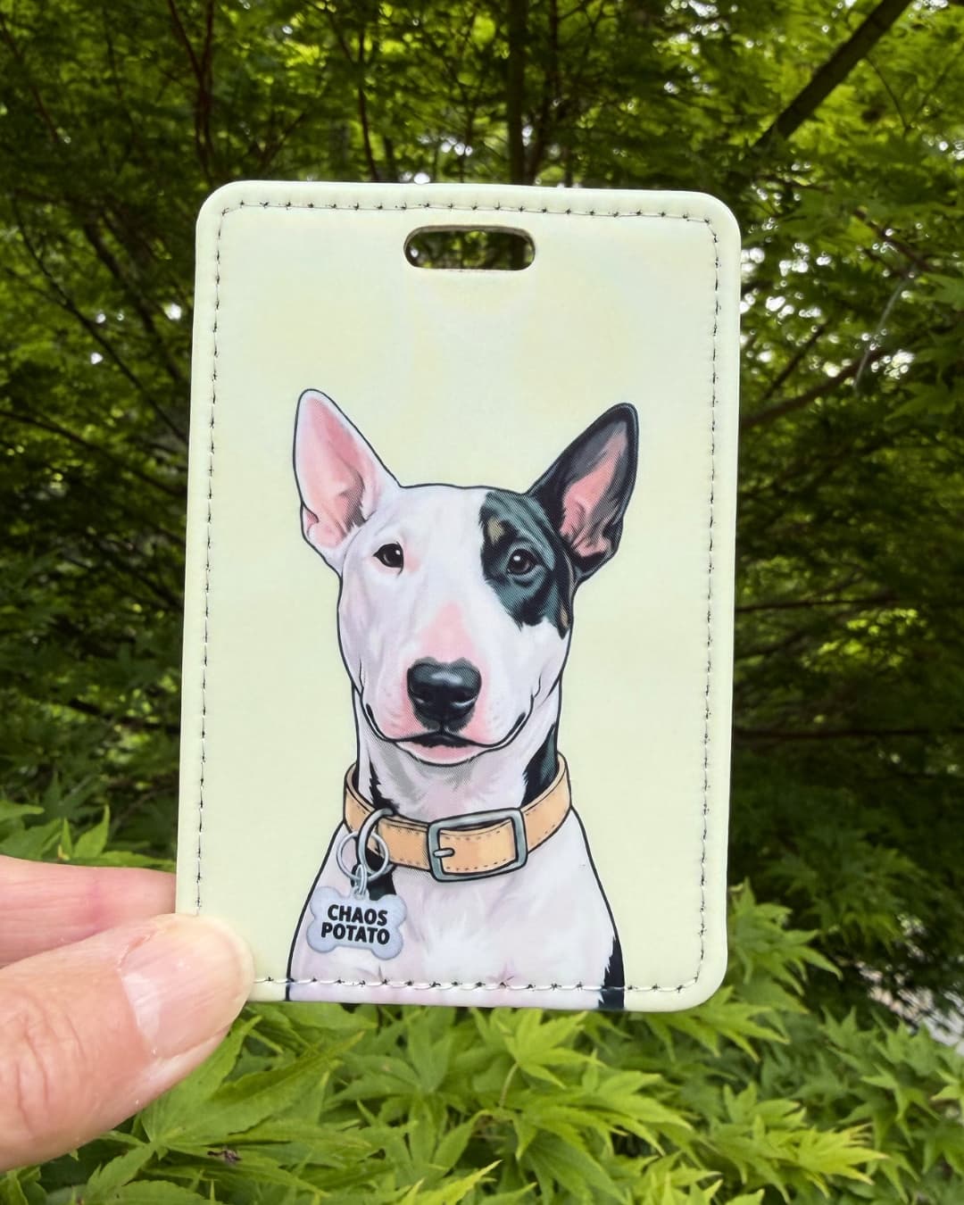 Bull Terrier luggage tag lifestyle; hand holding the faux leather Bull Terrier Chaos Potato luggage tag outdoors with green leaves in the background.