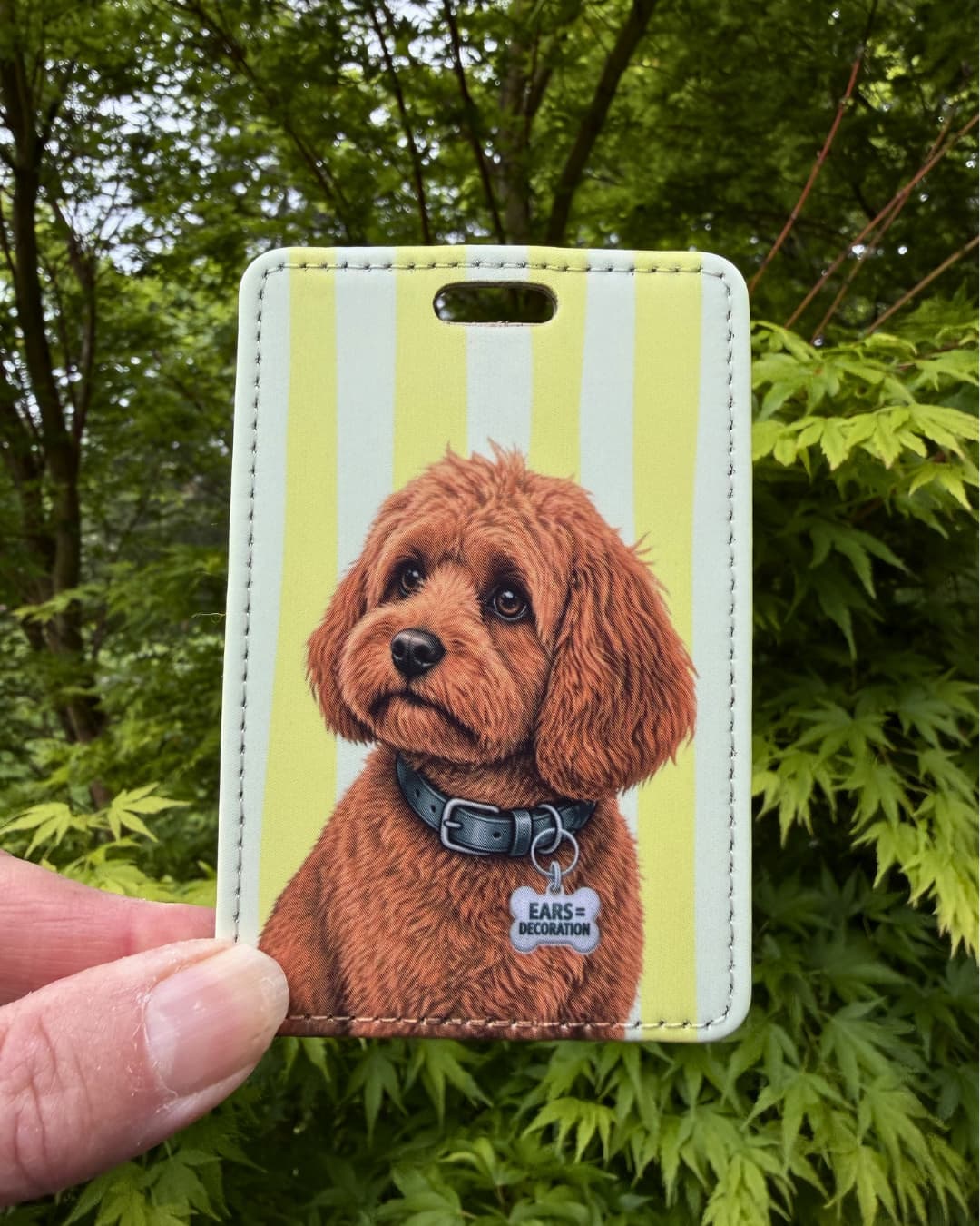 Cavapoo luggage tag lifestyle; Hand holding a fun faux leather luggage tag featuring an apricot curly-haired Cavapoo with an Ears Equal Decoration charm, vertical yellow and white stripes, green tree foliage background.