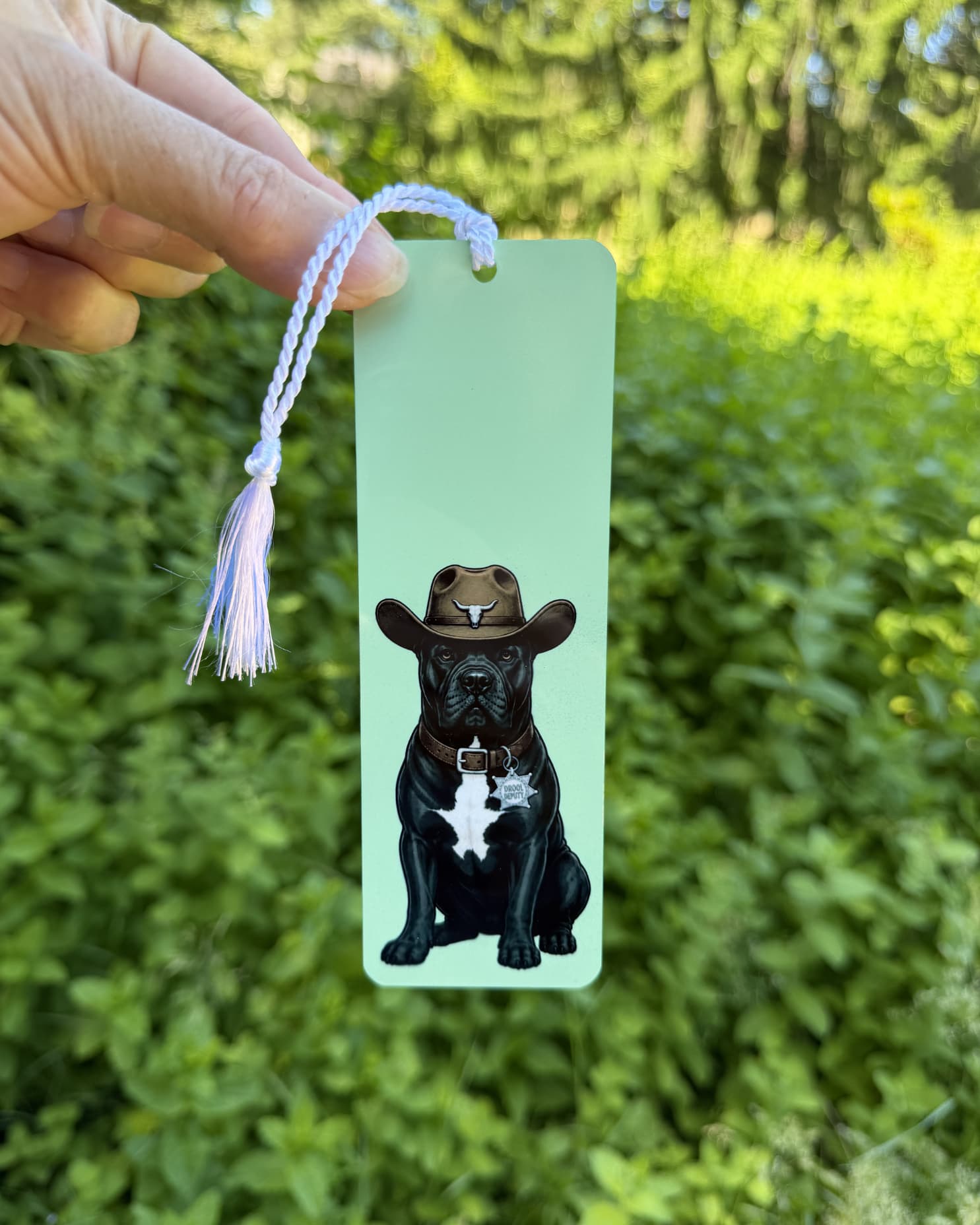 Hand holding cowboy Cane Corso bookmark against green foliage, sage green aluminum, Drool Deputy collar tag, white tassel
