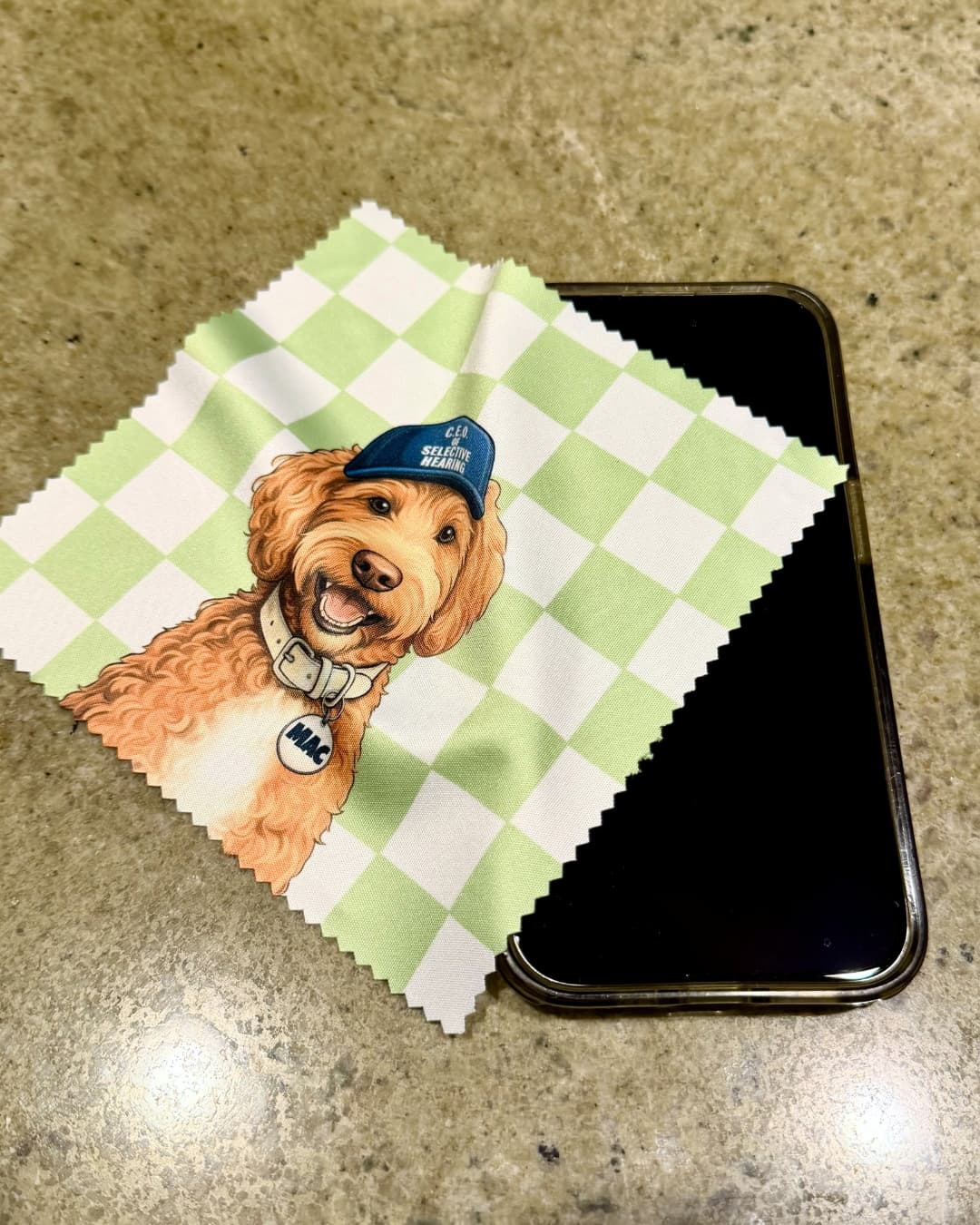 Custom microfiber cleaning cloth with goldendoodle pet art on green checkered background resting on a smartphone on a granite counter.