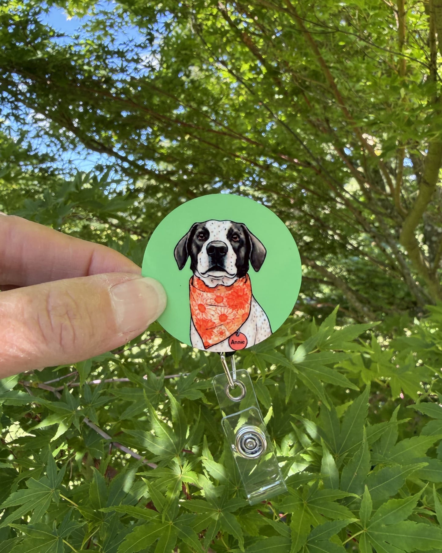 Custom dog badge reel lifestyle, hand holding retractable badge reel with illustrated black and white dog in orange bandana on green background, outdoors