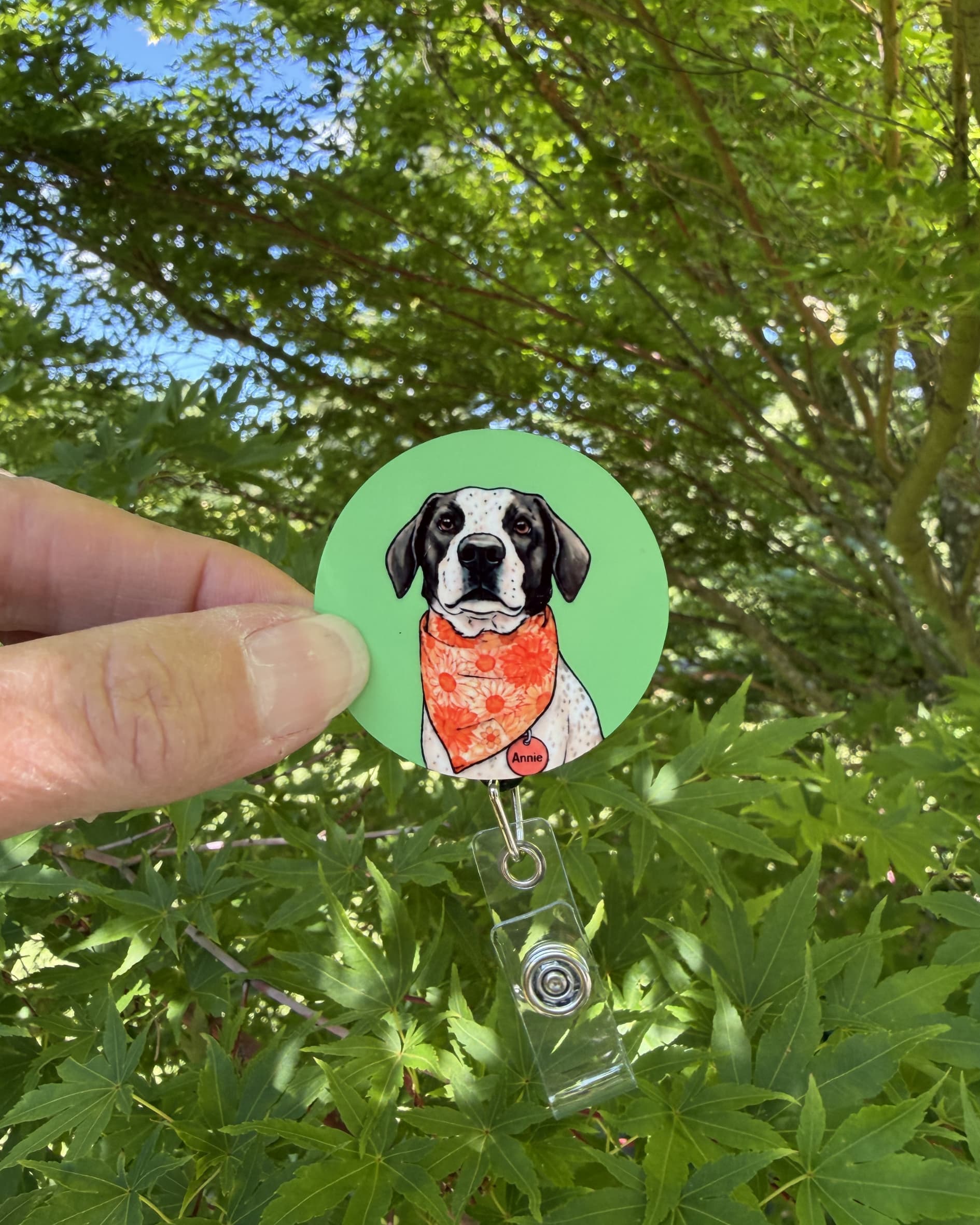 Custom dog badge reel lifestyle, hand holding retractable badge reel with illustrated black and white dog in orange bandana on green background, outdoors