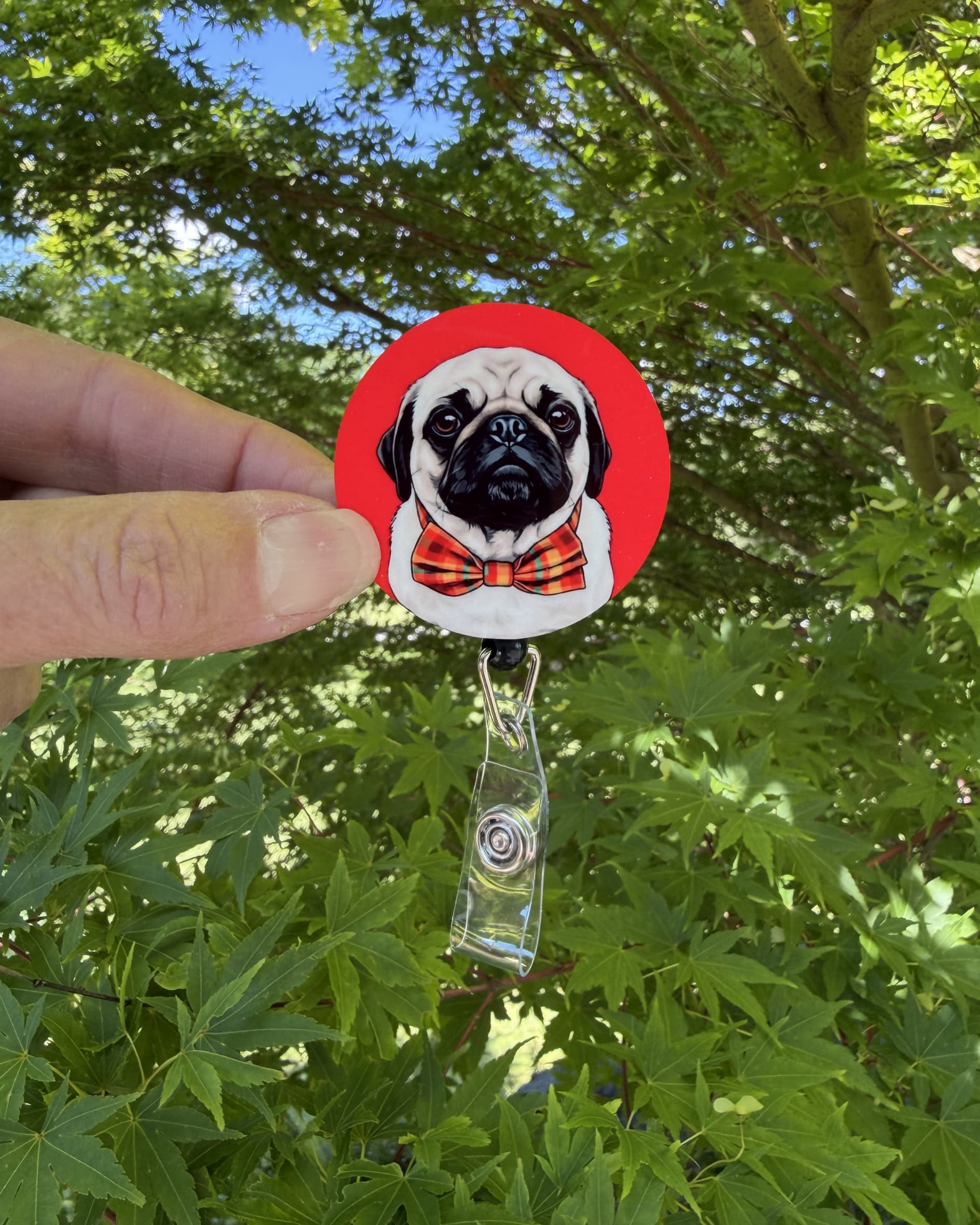 Custom pet badge reel lifestyle, hand holding retractable badge reel with illustrated pug in red plaid bowtie on red background, outdoors