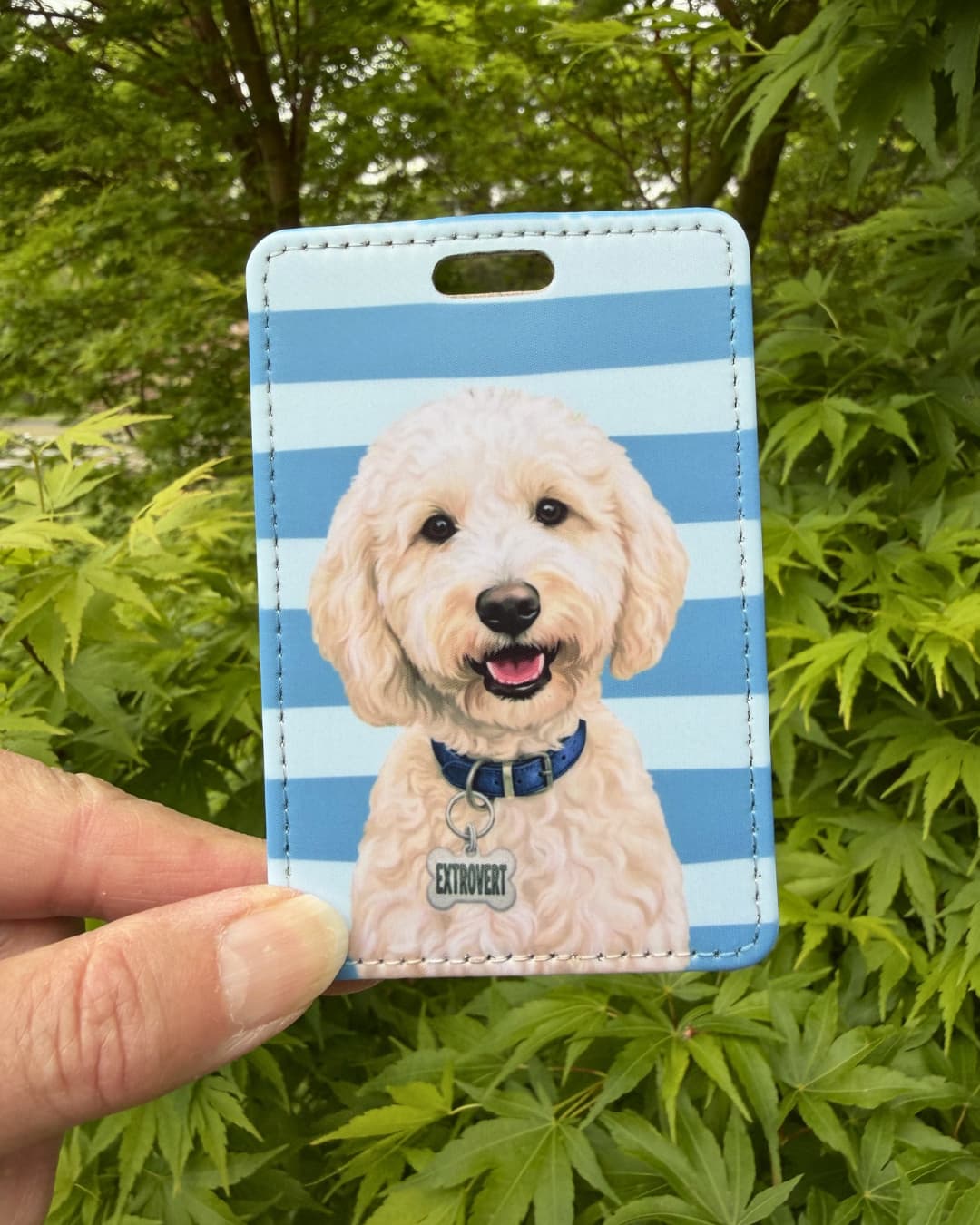 Cute doodle dog luggage tag lifestyle; Hand holding a fun faux leather luggage tag featuring a smiling cream Goldendoodle with a blue collar and Extrovert charm, light blue and white horizontal stripes, green tree foliage background.