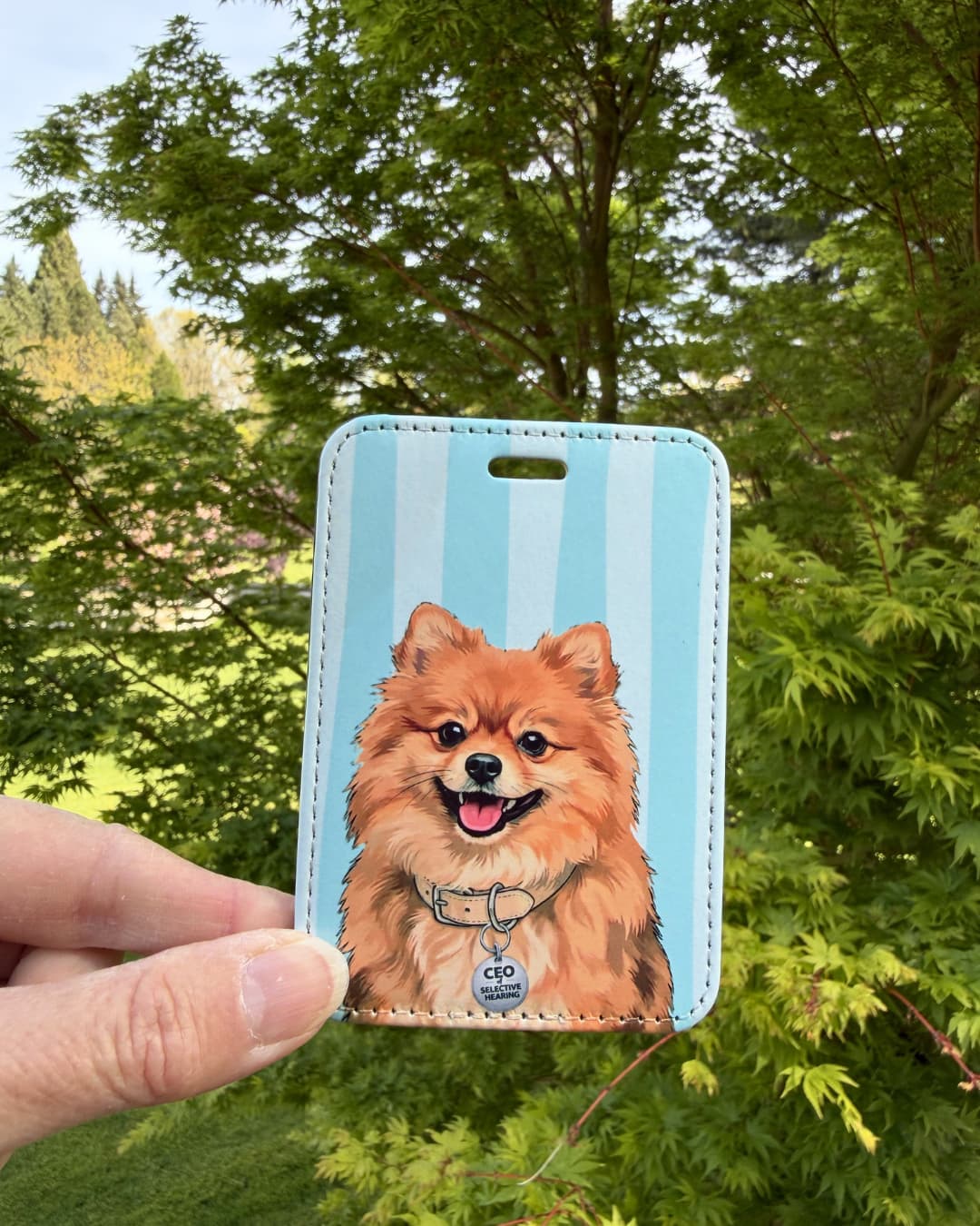 cute pomeranian luggage tag held outdoors against green trees showing smiling orange Pomeranian with CEO of Selective Hearing collar tag on blue and white striped faux leather tag