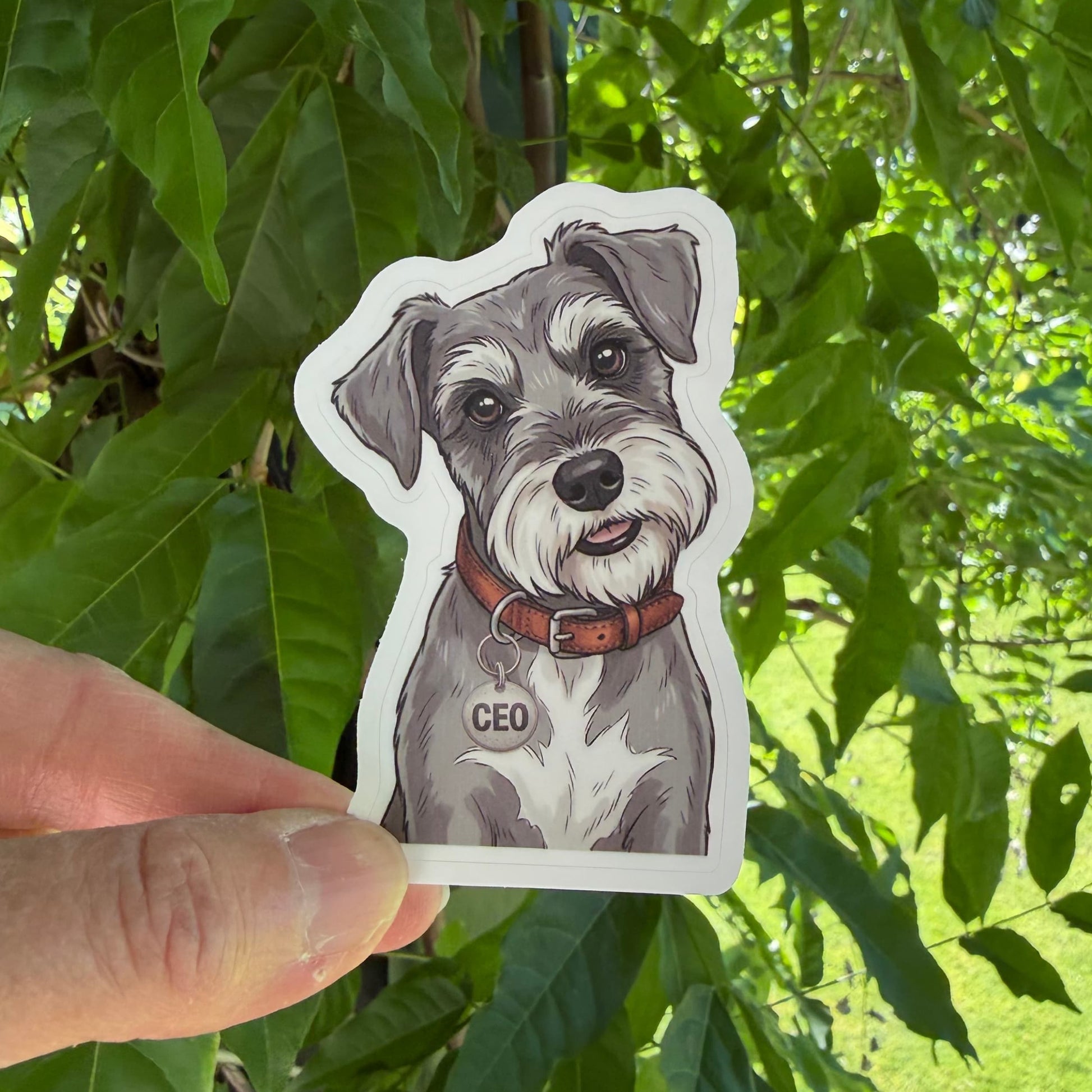 Cute Schnauzer sticker held in one hand against leafy green trees, showing the illustrated salt-and-pepper Schnauzer portrait and white kiss-cut border.
