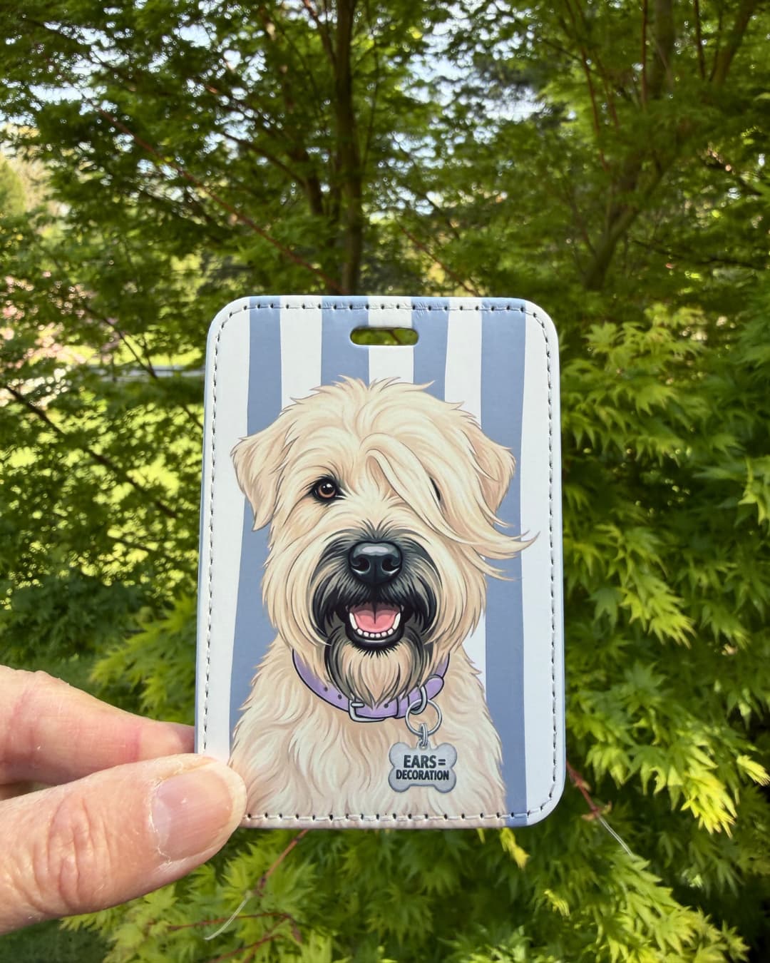 Cute wheaten terrier luggage tag lifestyle; Hand holding a fun faux leather luggage tag featuring a fluffy honey-colored Wheaten Terrier with a purple collar and Ears Equal Decoration charm, vertical light blue and white stripes, green tree foliage background.