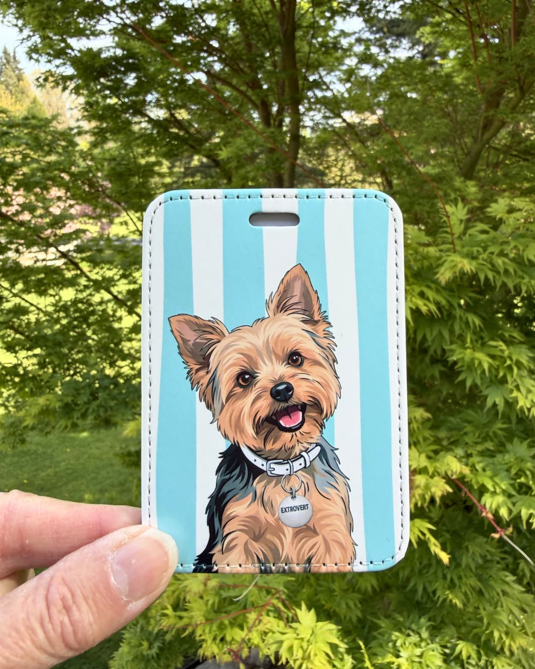 Cute Yorkie luggage tag lifestyle; Hand holding a fun faux leather luggage tag featuring a tan and black Yorkshire Terrier with a white collar and E"xtrovert" tag, vertical light blue and white stripes, green tree foliage background.