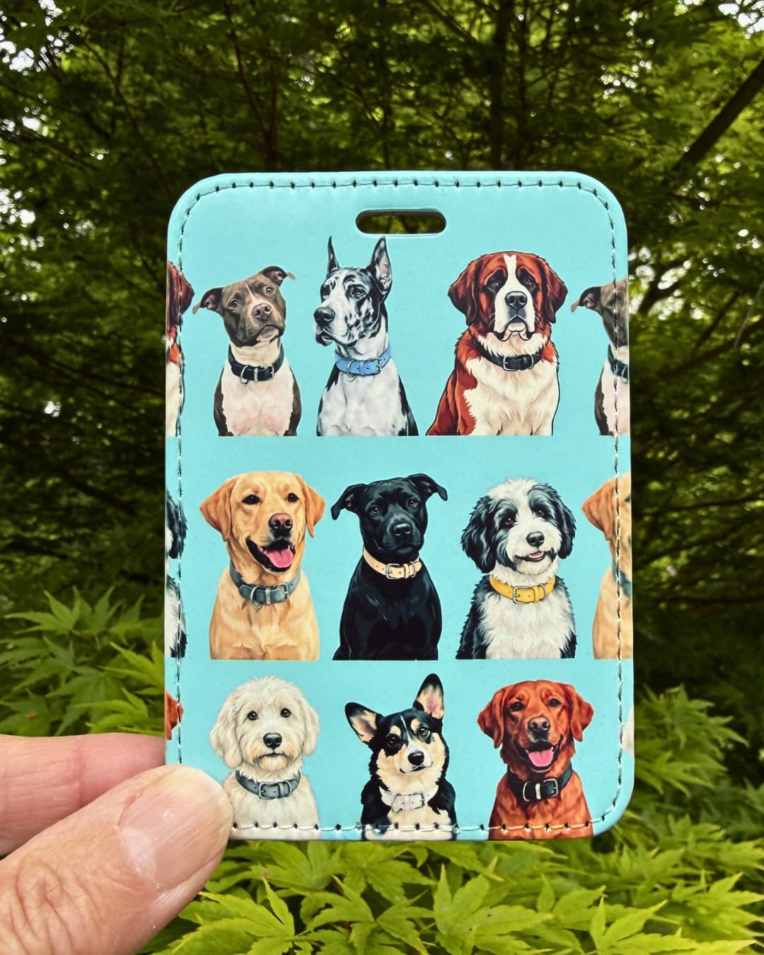 Dog lover luggage tag lifestyle; hand holding a teal faux leather multi-breed dog luggage tag outdoors with green Japanese Maple leaves in the background.