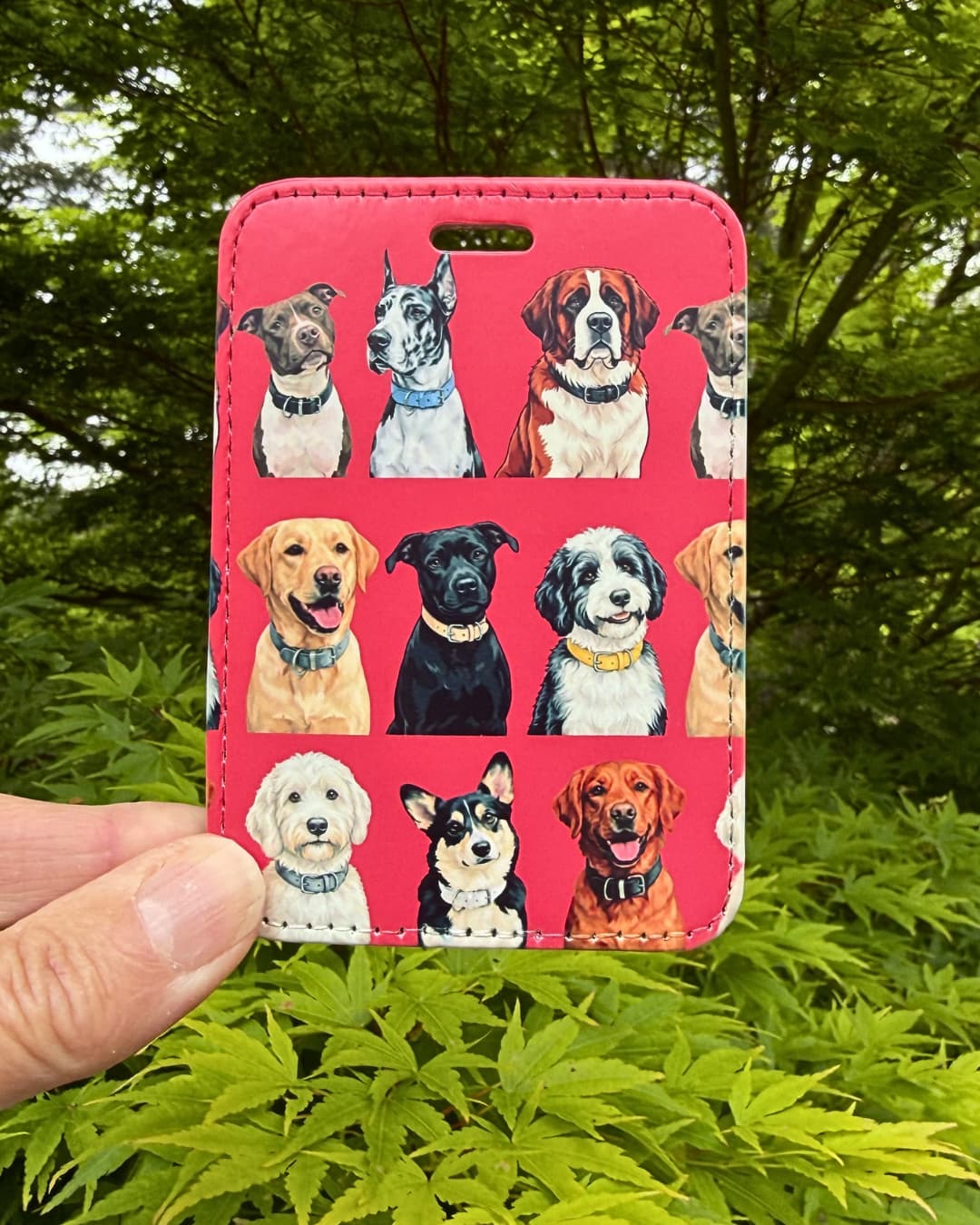 Dog lover luggage tag lifestyle; hand holding a red faux leather multi-breed dog luggage tag outdoors with green Japanese Maple leaves in the background.