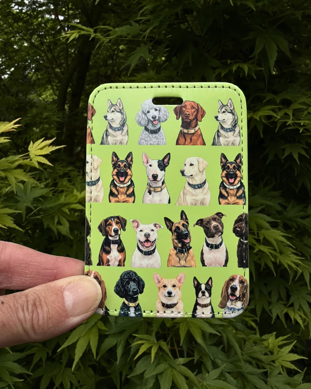 Dog lover travel gift lifestyle green; hand holding a green faux leather multi-breed dog luggage tag outdoors with green Japanese Maple leaves in the background.