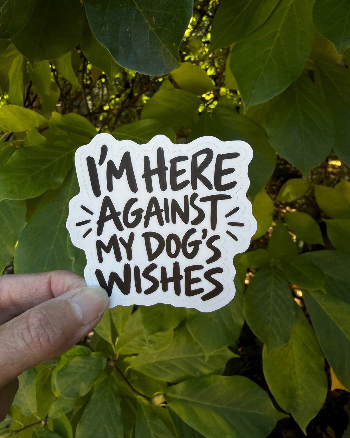 Dog mom humor sticker held between fingers against green leaves, reading “I’M HERE AGAINST MY DOG’S WISHES.