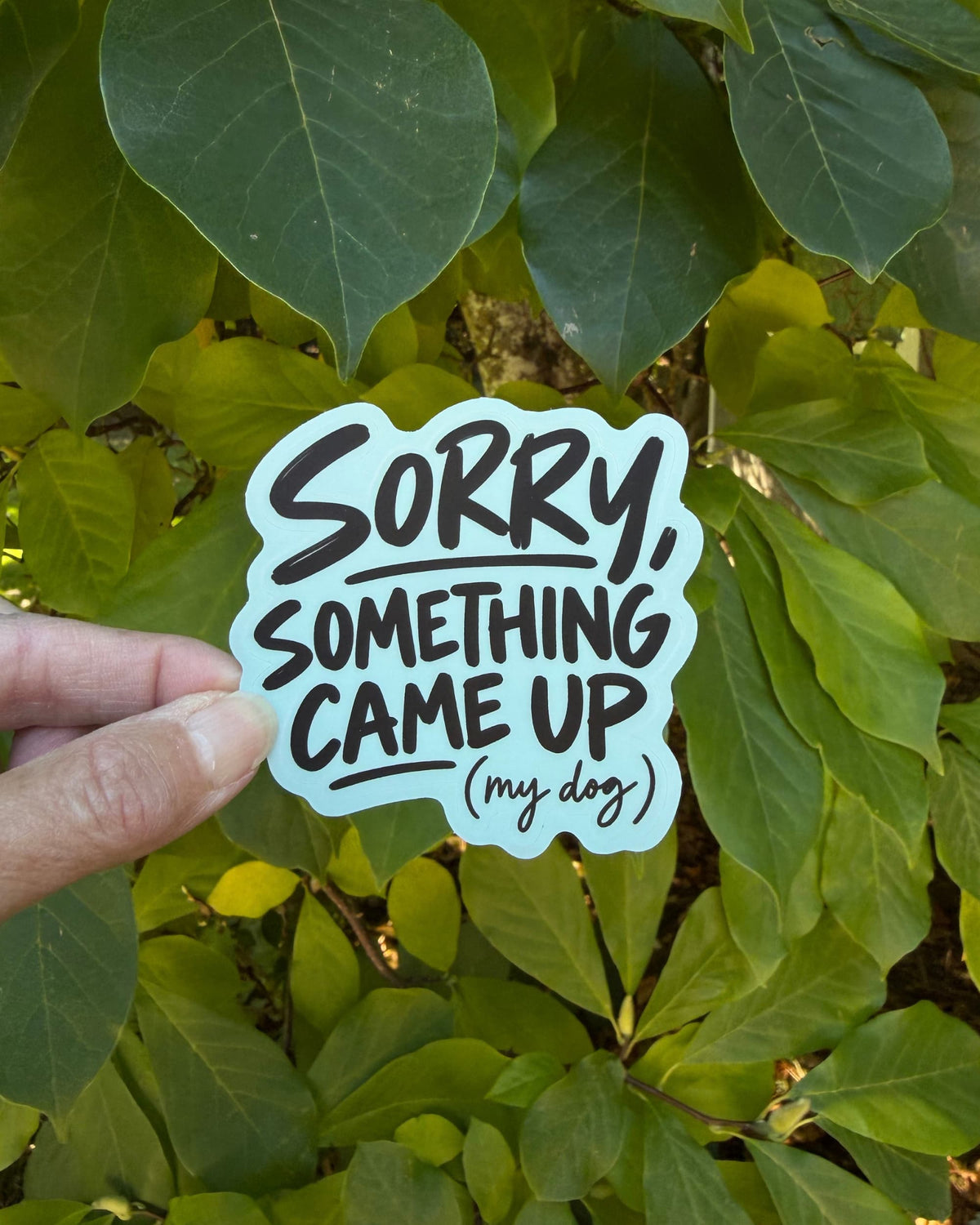 dog parent humor sticker with black lettering and a light blue sticker background, reading “SORRY, SOMETHING CAME UP (my dog),” held between fingers in front of green leaves.