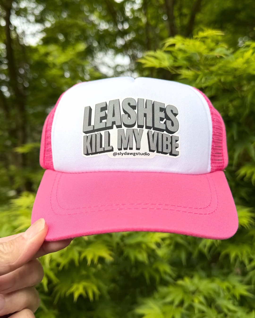 Dog trucker hat with saying lifestyle pink; pink and white Leashes Kill My Vibe dog trucker hat held outdoors with green foliage in the background.
