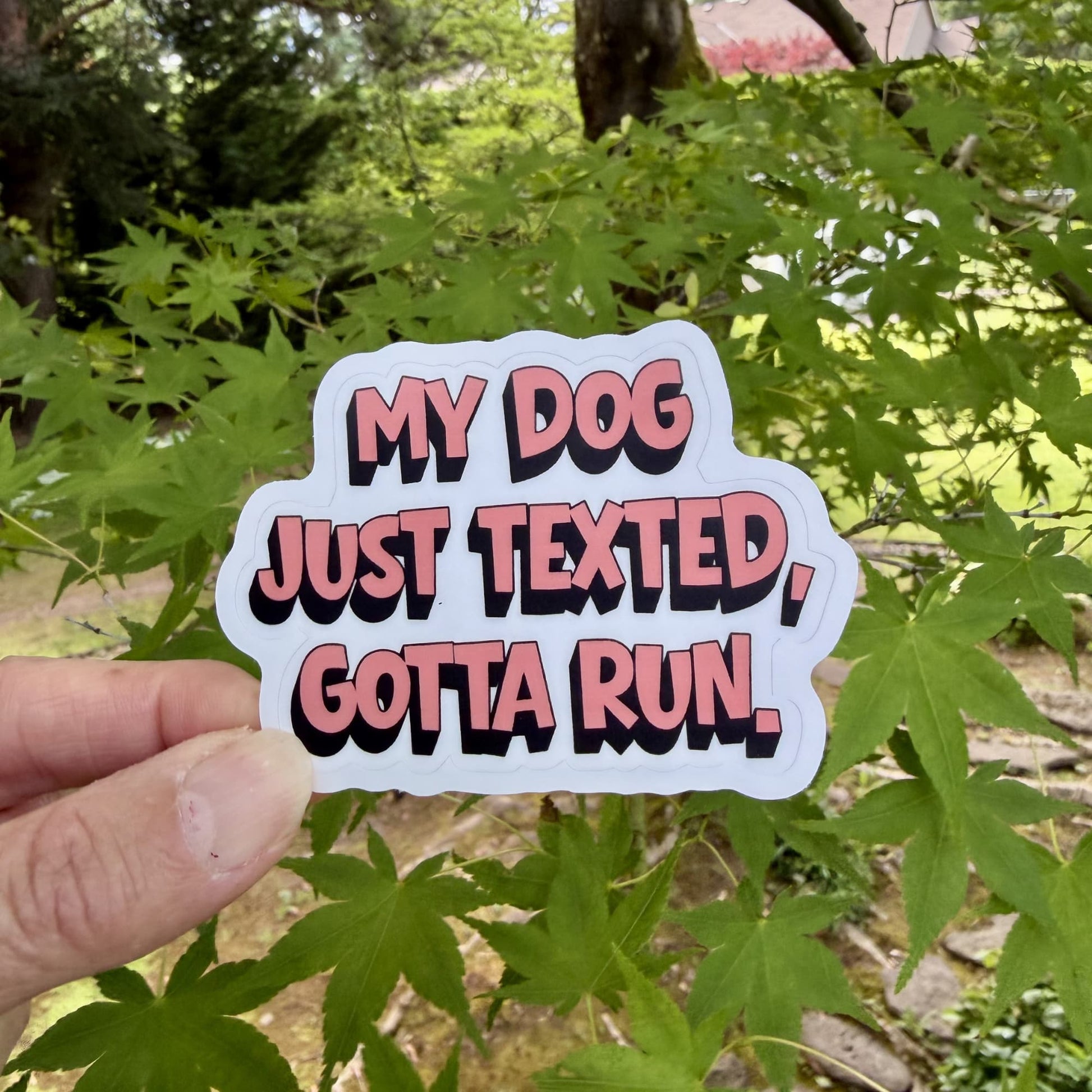 Hand holding My Dog Just Texted Gotta Run funny dogs before people sticker outdoors against green leaves.