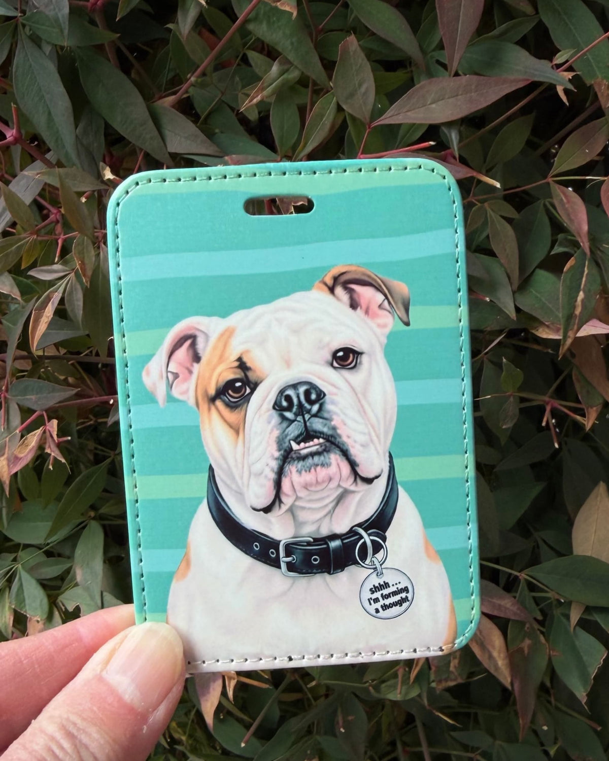Hand holding English Bulldog luggage tag, white bulldog with fawn eye patch on teal stripes, charm reads shhh... I'm forming a thought