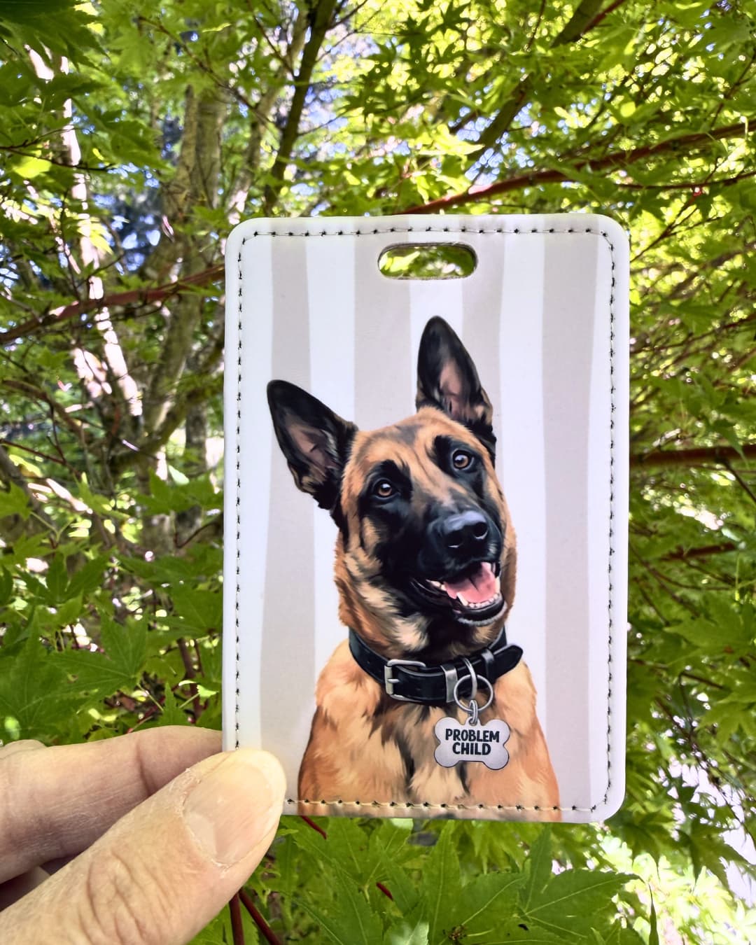 Funny Belgian Malinois luggage tag lifestyle; Hand holding a faux leather luggage tag featuring a grinning Belgian Malinois with a Problem Child funny bone charm, tan and white stripes, green tree foliage background.
