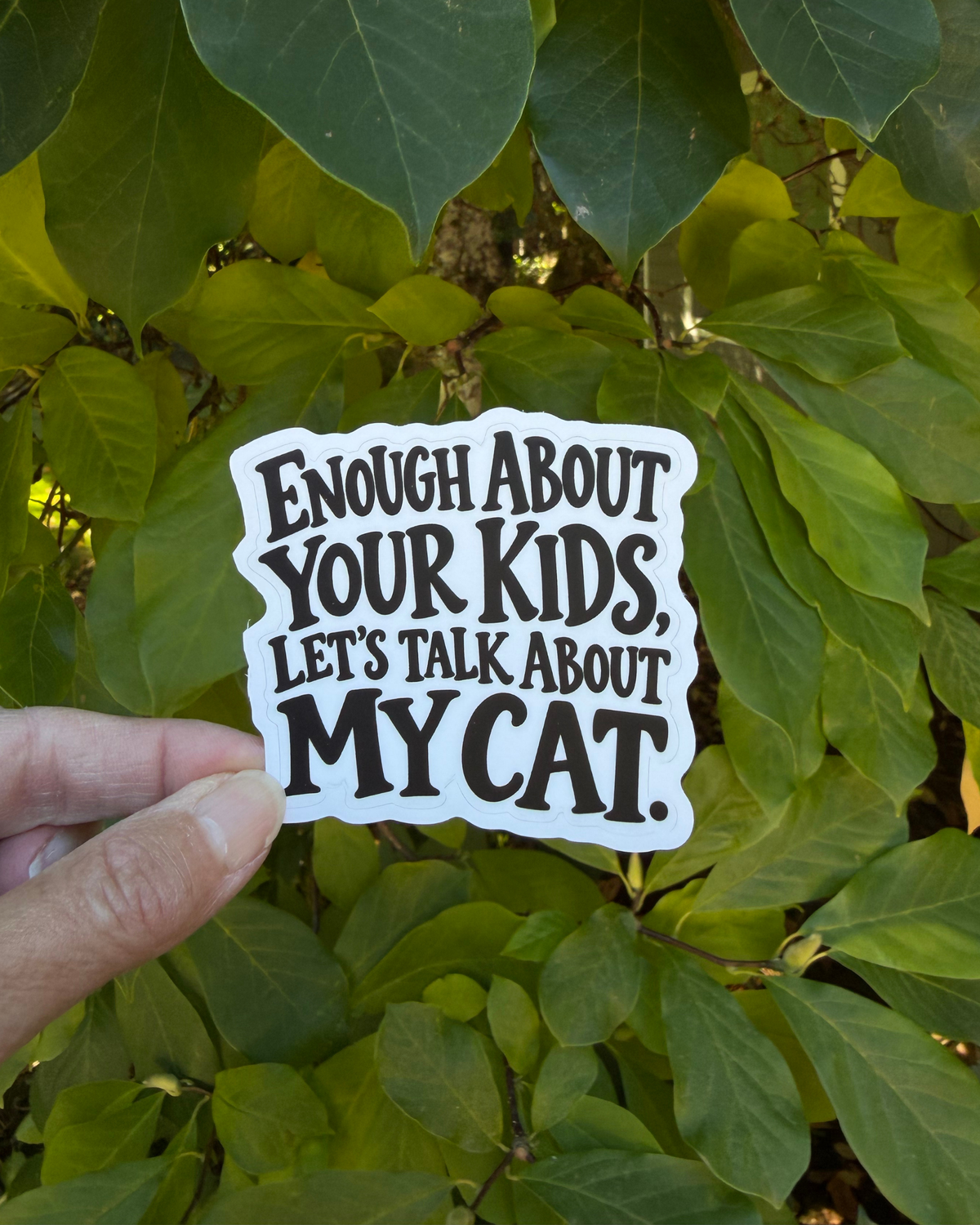 Funny cat owner sticker with black lettering on a white background reading “Enough About Your Kids, Let’s Talk About My Cat,” held between fingers against green leaves.
