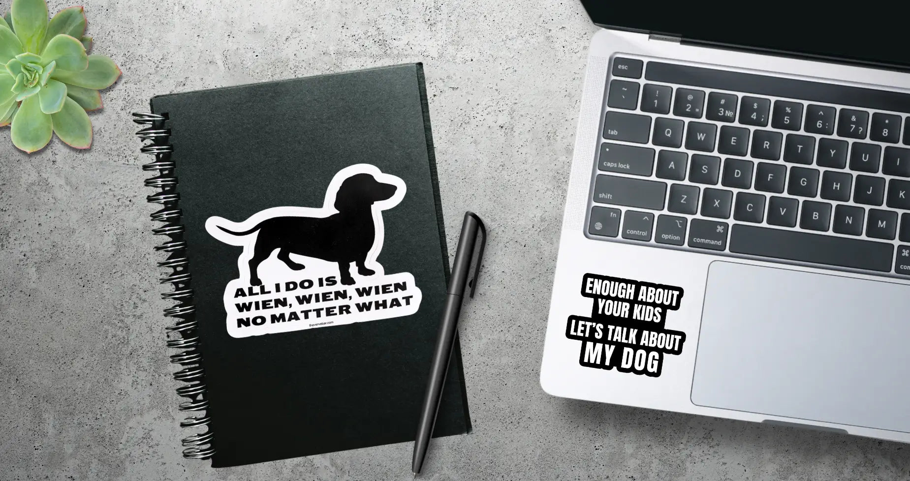 Black notebook with funny dog sticker: dachshund illustration and text next to a laptop with a dog parent sticker. All sitting on concrete surface.