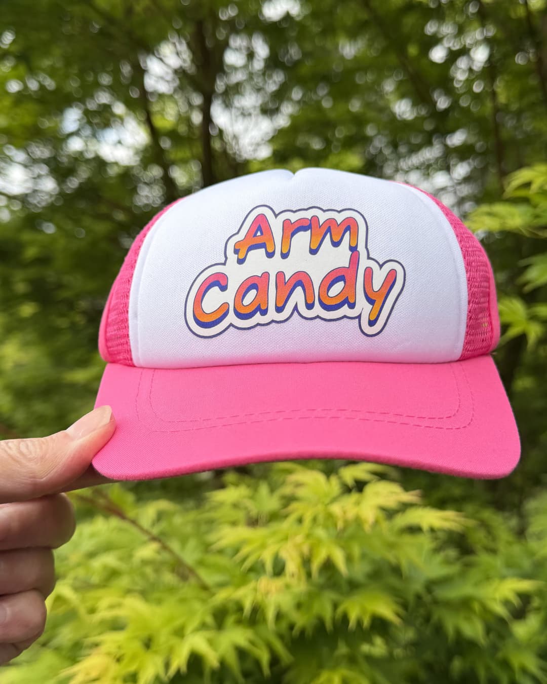 Funny dog trucker hat lifestyle; hand holding a hot pink and white Arm Candy dog trucker hat outdoors with green Japanese Maple leaves in the background.