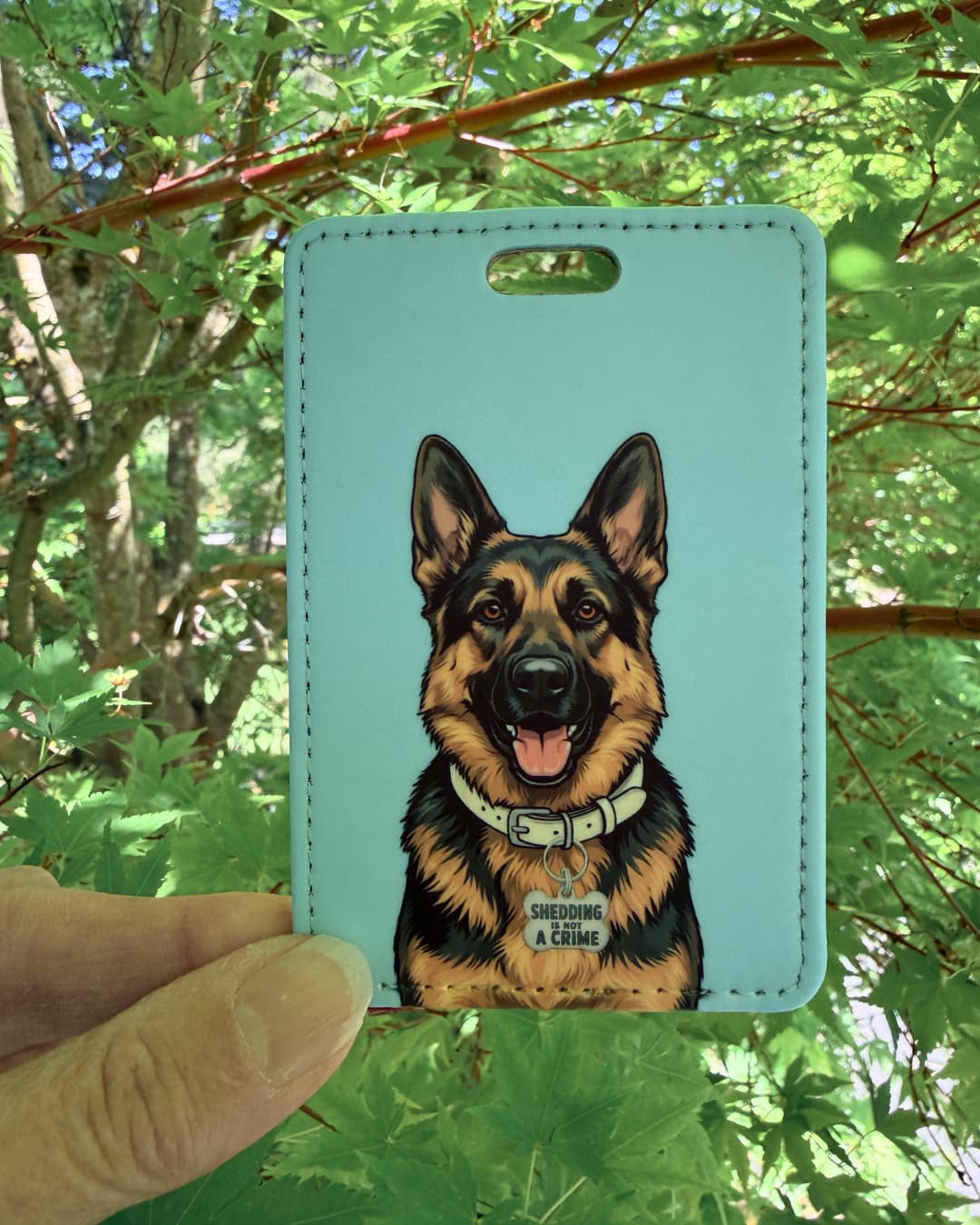 Funny German Shepherd luggage tag lifestyle; Hand holding a fun faux leather luggage tag featuring a wide-eyed grinning German Shepherd with a Problem Child collar charm, light blue and white background, green tree foliage background.