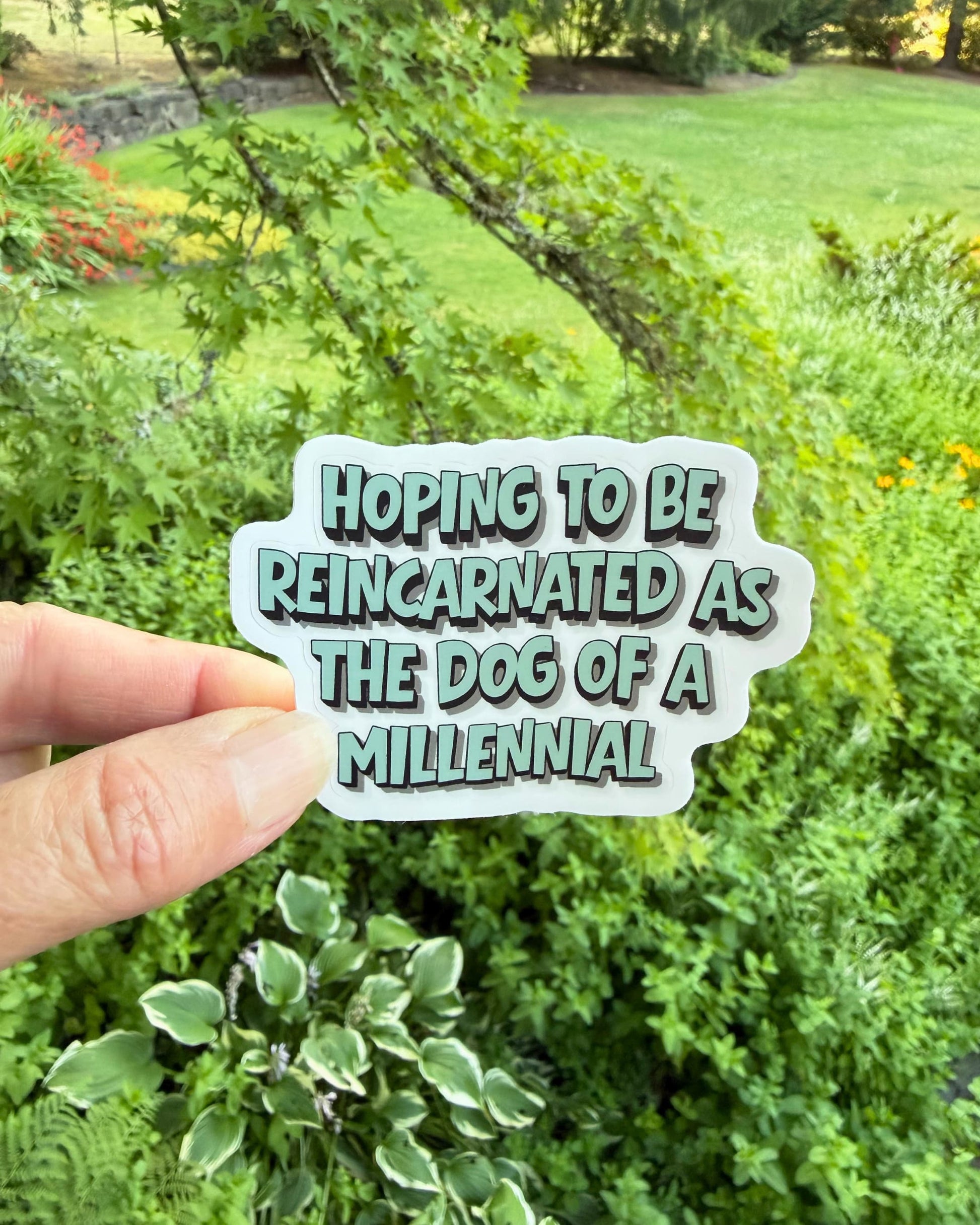 Funny millennial dog parent sticker held in one hand in front of leafy green trees and garden foliage.
