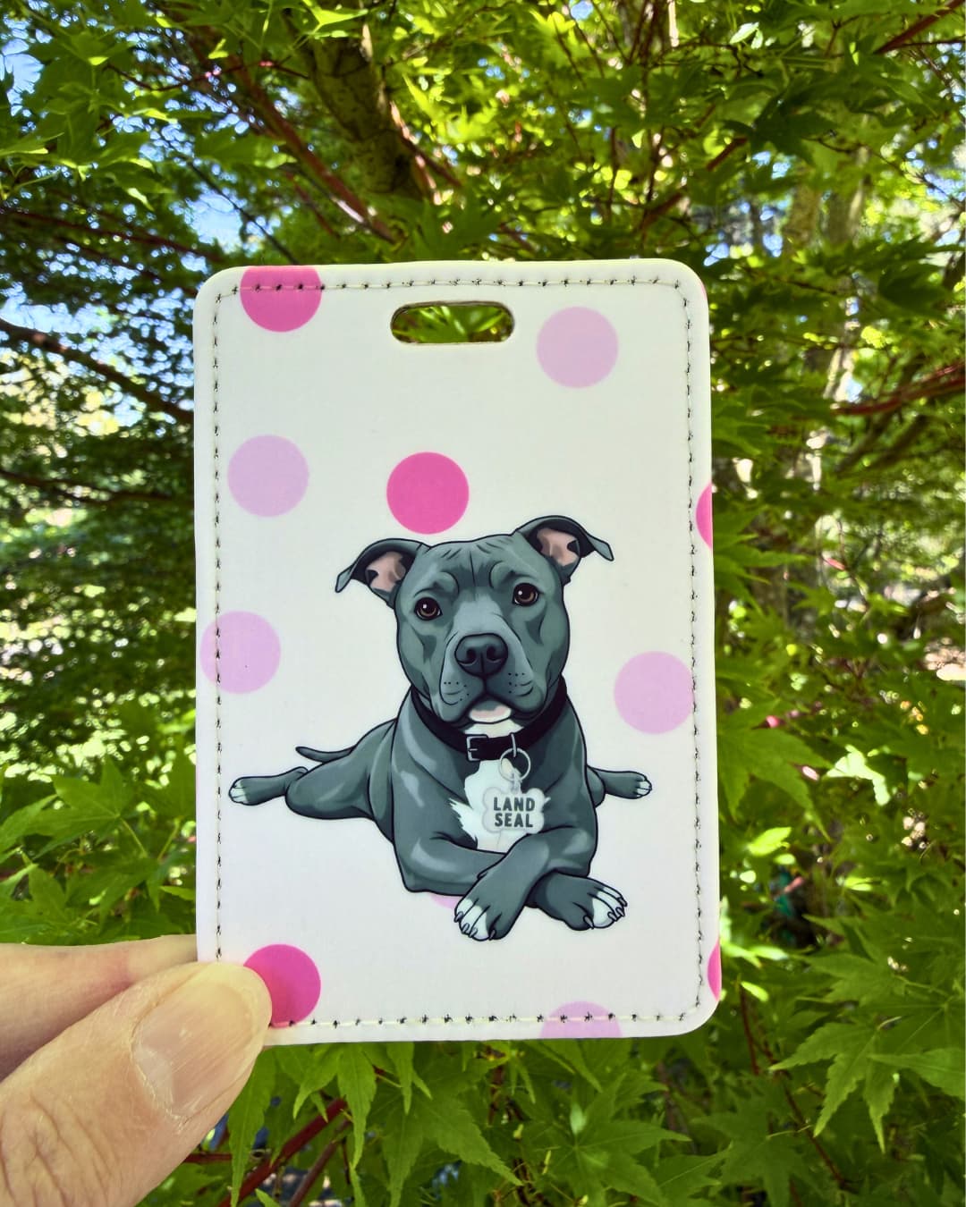 Funny pit bull luggage tag light pink polka dot lifestyle; Hand holding a fun faux leather luggage tag featuring a grey pit bull in sploot pose with Land Seal charm, light pink polka dot background, green tree foliage background.
