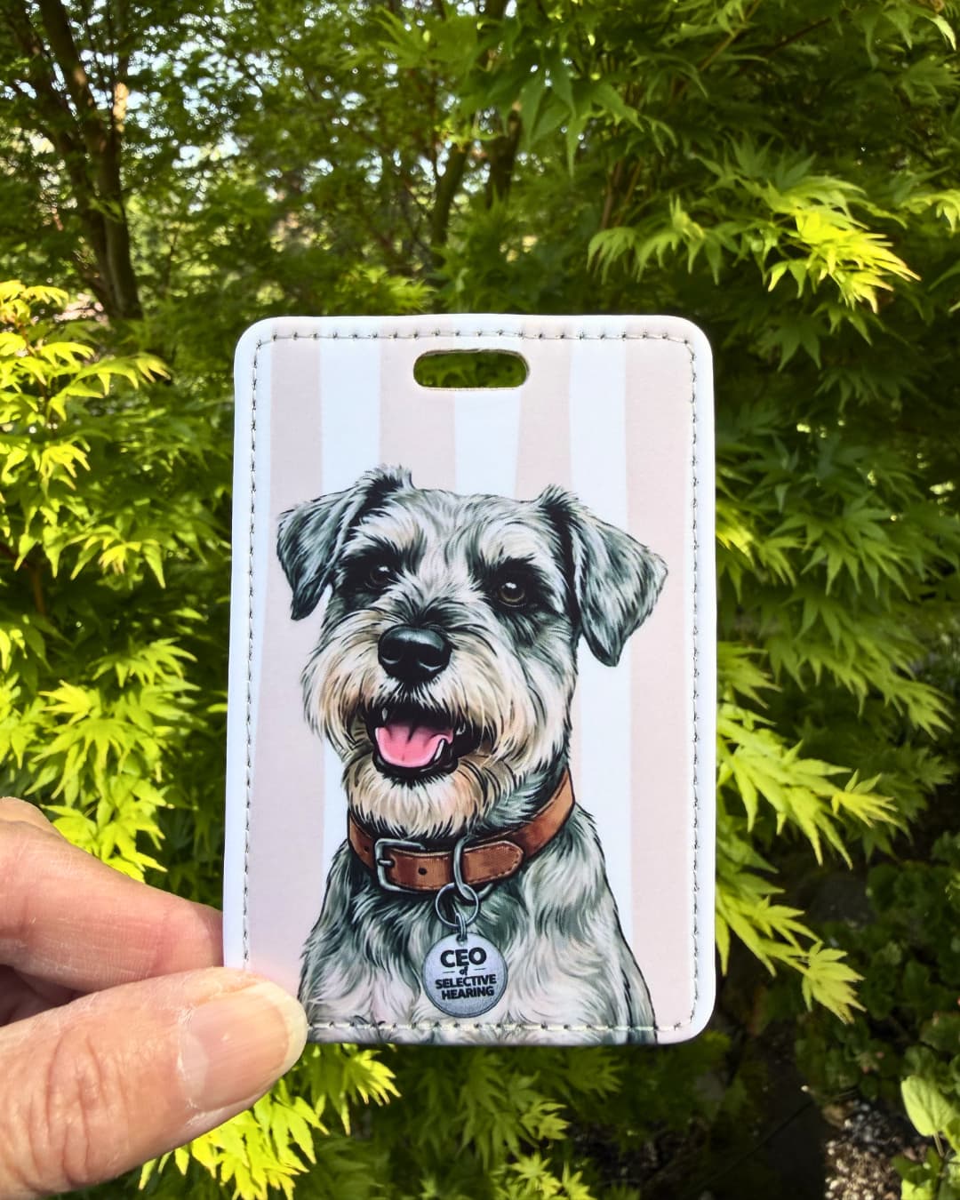 Funny schnauzer luggage tag lifestyle; Hand holding a fun faux leather luggage tag featuring a salt and pepper Schnauzer with a brown collar and CEO of Selective Hearing charm, vertical tan and white stripes, green tree foliage background.