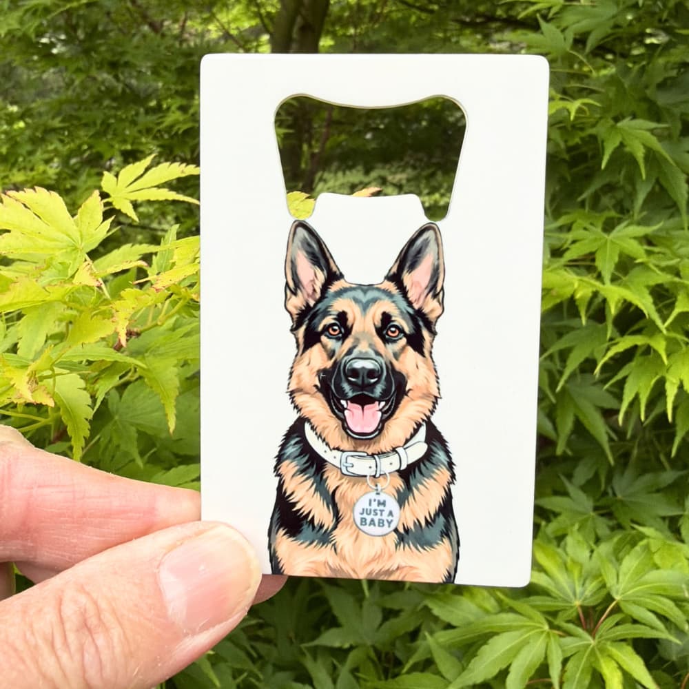 German Shepherd bottle opener lifestyle; a hand holding a flat metal GSD bottle opener outdoors with green leaves in the background.