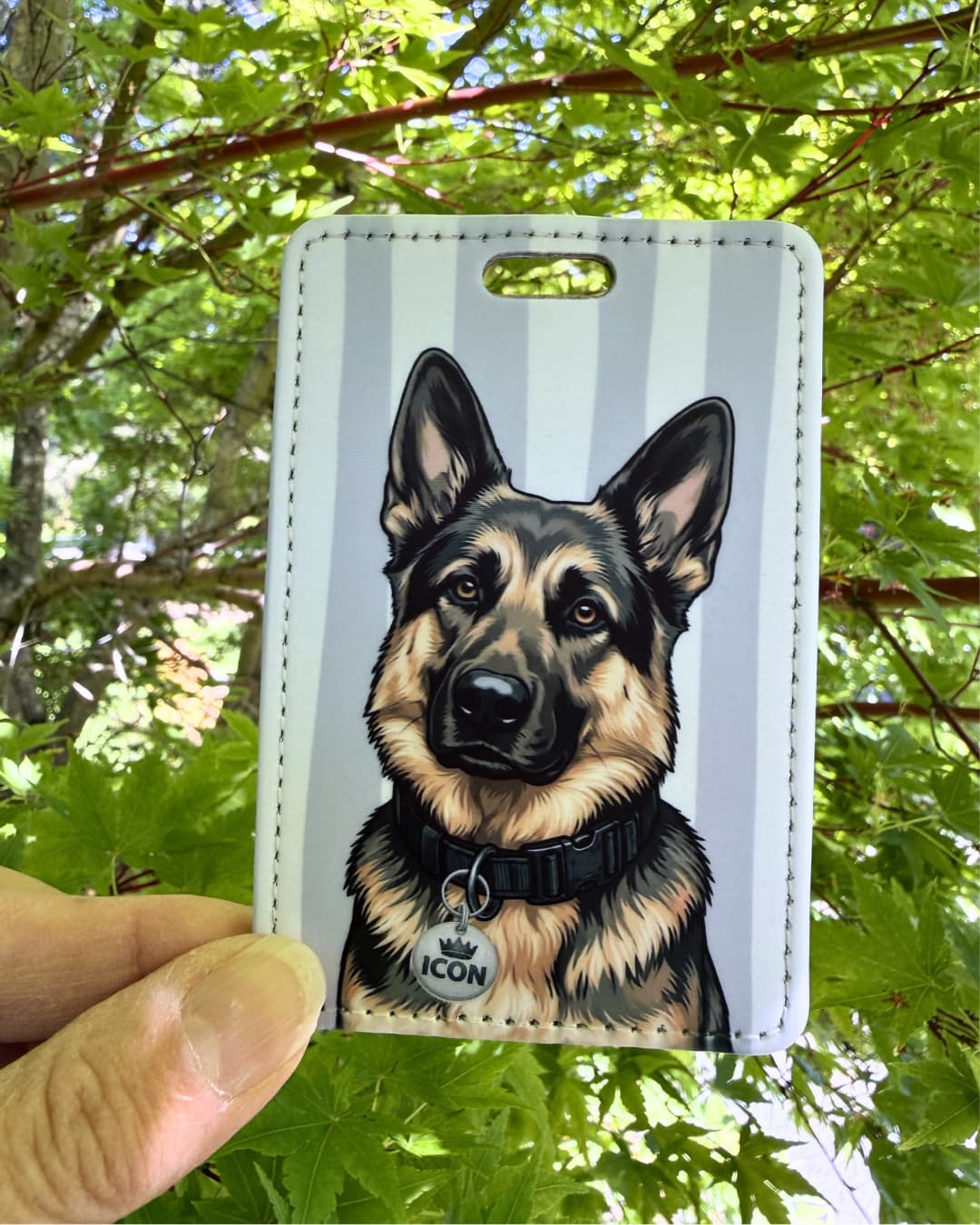German Shepherd luggage tag lifestyle; Hand holding a faux leather luggage tag featuring a regal German Shepherd with an Icon crown charm, grey and white stripes, green tree foliage background.