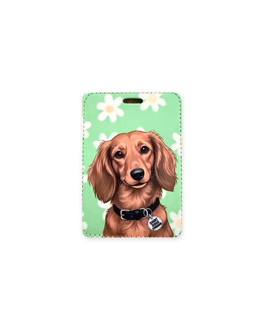 Long haired red dachshund luggage tag with an original design of a doxie with silky red ears, wearing a black collar and a round charm that reads "Short Dogg Syndrome", on mint green with white daisies. Faux leather luggage tag with stitched edges and a slot at the top.