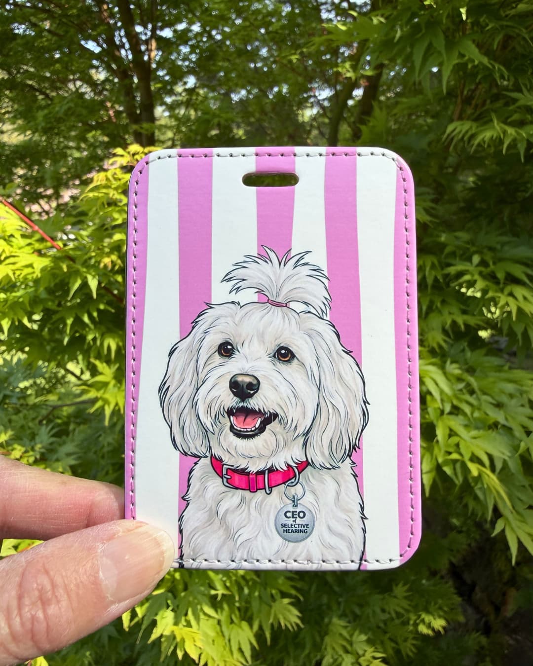 Maltese luggage tag lifestyle; Hand holding a vibrant faux leather luggage tag featuring a groomed white Maltese with a pink bow and CEO of Selective Hearing collar charm, pink and white stripes, green tree foliage background.
